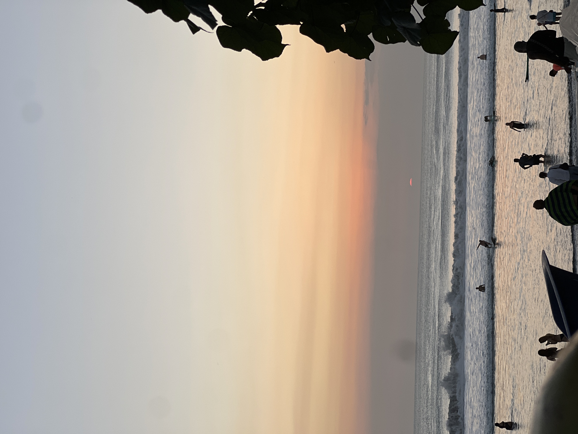 In Kuta, Indonesia, a serene beach scene unfolds at sunset. The sky transitions from a pale blue at the zenith to soft oranges and pinks near the horizon, where a sliver of the sun is visible just above the sea. Gentle waves break on the shore, creating a foamy expanse of water that reflects the muted colors of the sky. A surfer rides a wave in the mid-ground, while other figures are scattered throughout the shallow water, some wading and others standing. In the foreground, a few people are gathered on the wet sand, with a dark, boat-like object partially obscuring the lower left corner. The overall atmosphere is tranquil and captures a typical evening on Kuta Beach.