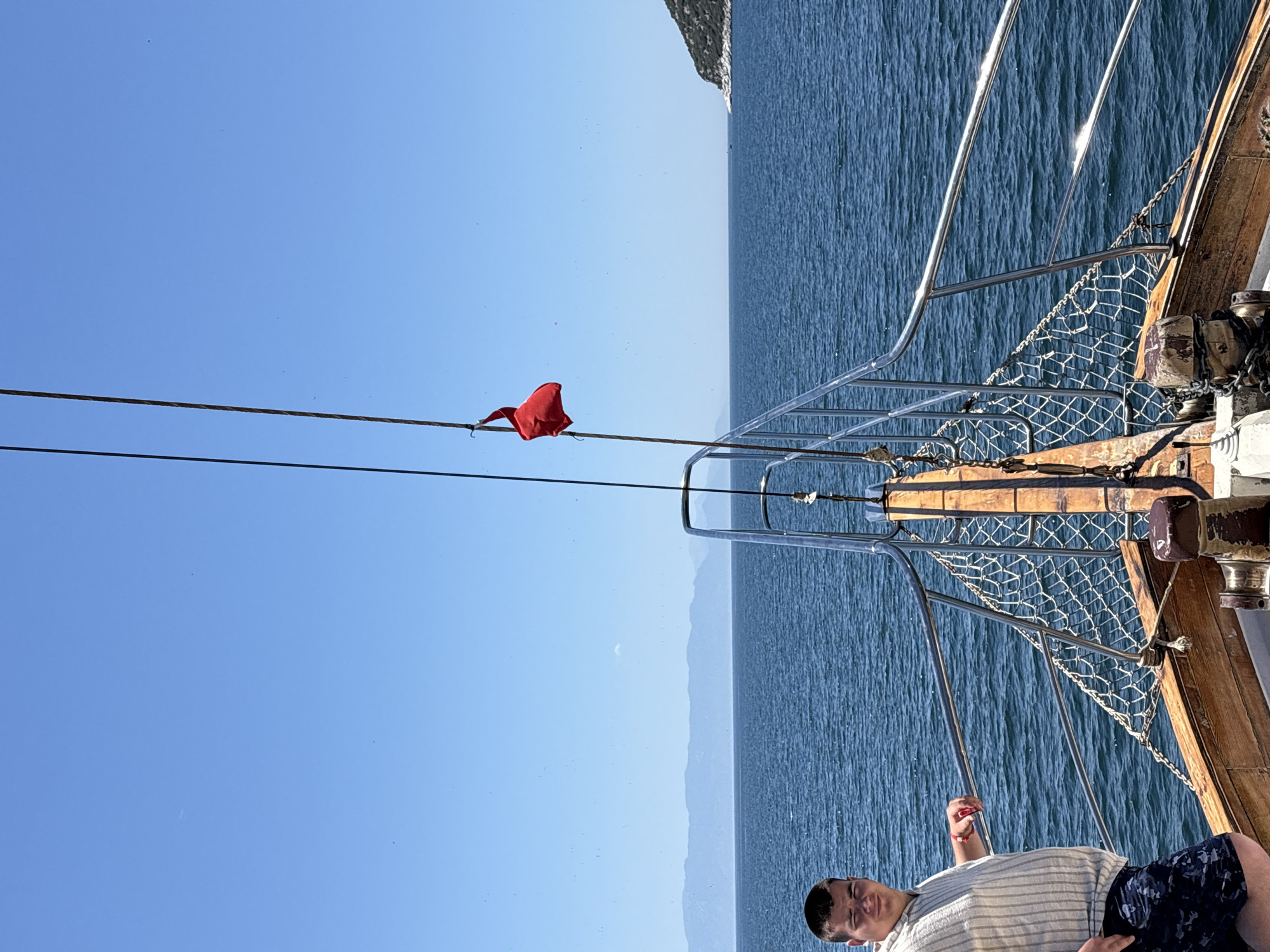 The image captures a sunny daytime scene aboard a wooden boat, likely cruising on a body of water in Menderes, Turkey. In the lower right foreground, a young man is visible, lying down and appearing relaxed, wearing a light-colored striped shirt and dark patterned shorts. His eyes are partially closed, possibly due to the bright sunlight.
The boat features traditional wooden decking, sturdy metallic railings, and a protective netting along the side. A red cloth, resembling a small flag, is tied to a rope or mast extending from the boat's structure on the left side of the frame. The vast, rippling blue sea stretches towards the horizon, where distant, hazy mountains are visible under a clear, bright blue sky.
The weather is distinctly sunny and clear, with strong sunlight creating shimmering reflections on the water and the boat's metalwork. The overall atmosphere is one of leisure and relaxation, typical of a pleasant boat trip. No visible text is present in the image.