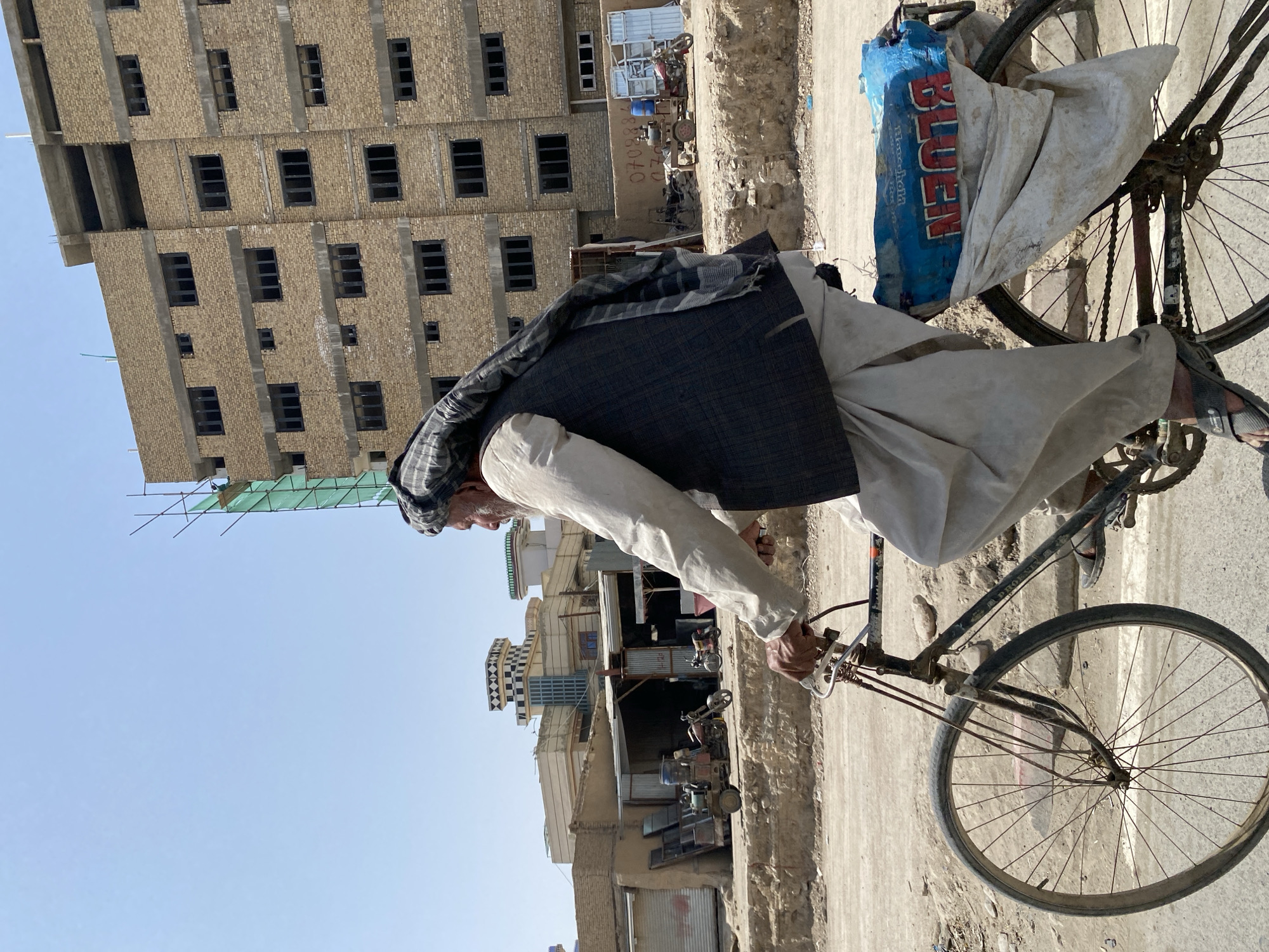 A man wearing traditional clothing, including a turban and vest, rides a bicycle through a dusty urban environment in Kandahar, Afghanistan. The bicycle is loaded with a blue tarp and what appears to be a white bag, possibly containing goods. In the background, a multi-story brick building under construction, featuring numerous dark, empty window frames, dominates the scene. Adjacent to this building are smaller, older structures, some with open storefronts, and a few motorcycles and a three-wheeled vehicle are visible. The ground is unpaved and appears to be a dirt road. The sky is clear and blue, indicating a sunny day.