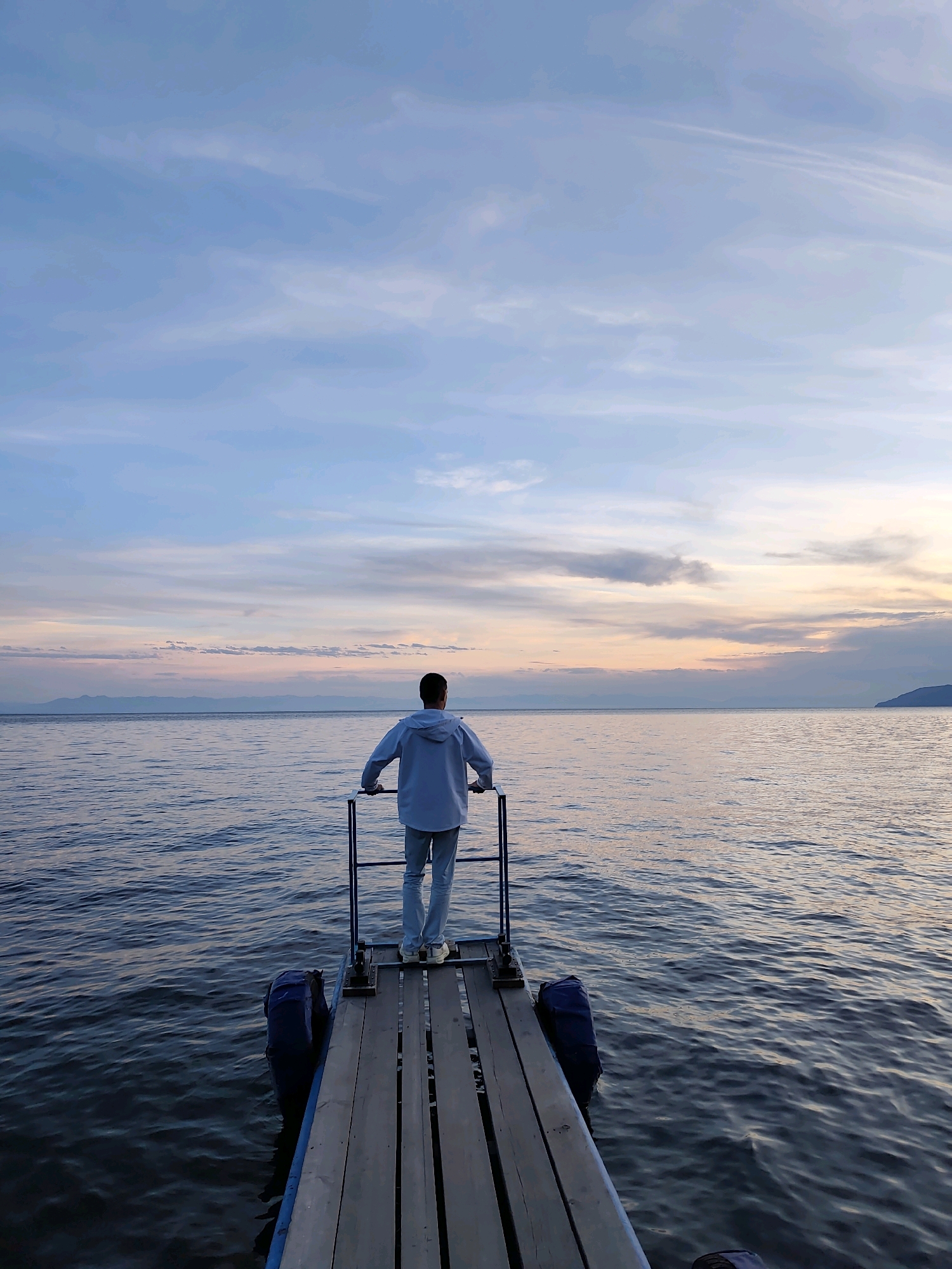 A person stands on a wooden pier extending into a large body of water. The person is facing away from the camera, looking out at the horizon. They are wearing a light blue hooded jacket and light blue jeans. The pier is made of weathered wooden planks and has a metal railing. Blue buoys are attached to the side of the pier. The water is calm with gentle ripples, reflecting the soft colors of the sky. The sky is a mix of blues, pinks, and oranges, indicating either sunrise or sunset. Distant mountains or hills can be seen along the horizon. The scene suggests a tranquil moment by the water, possibly on Lake Baikal in Listvyanka, Russia, given the characteristic landscape and the impression of a vast freshwater lake.