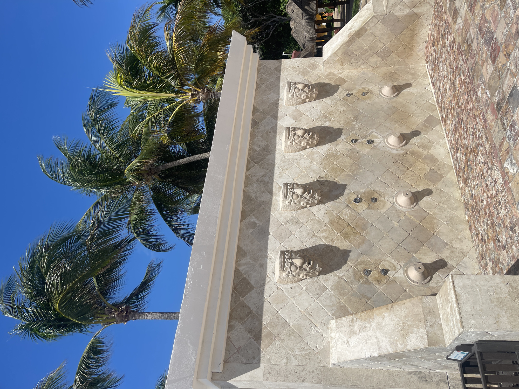 The image shows an outdoor shower structure in Playa del Carmen, Mexico. The structure is made of light-colored stone tiles arranged in a diamond pattern. The shower wall features four decorative sculpted elements, resembling ornate brackets, positioned vertically along the wall. Adjacent to each bracket are shower nozzles. A cream-colored wall extends upwards to the left of the shower wall. Palm trees are visible against a clear blue sky. The structure adjoins a wall made of small red and brown stones.