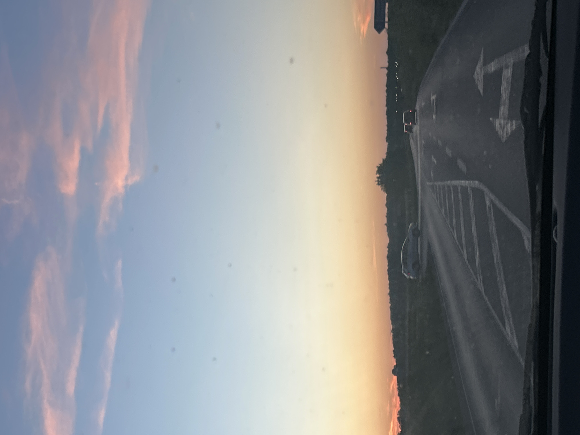 The image depicts a road scene at dusk. The sky is a gradient of pale blue and orange, with wispy pink clouds. A two-lane road curves into the distance, marked with white directional arrows and lines. Two cars are visible on the road, one in the distance and another further ahead. To the left of the road, there is a grassy embankment and some trees. The foreground shows the dashboard of a vehicle, indicating the photo was taken from inside a car. The scene is located in Unknown, Unknown.