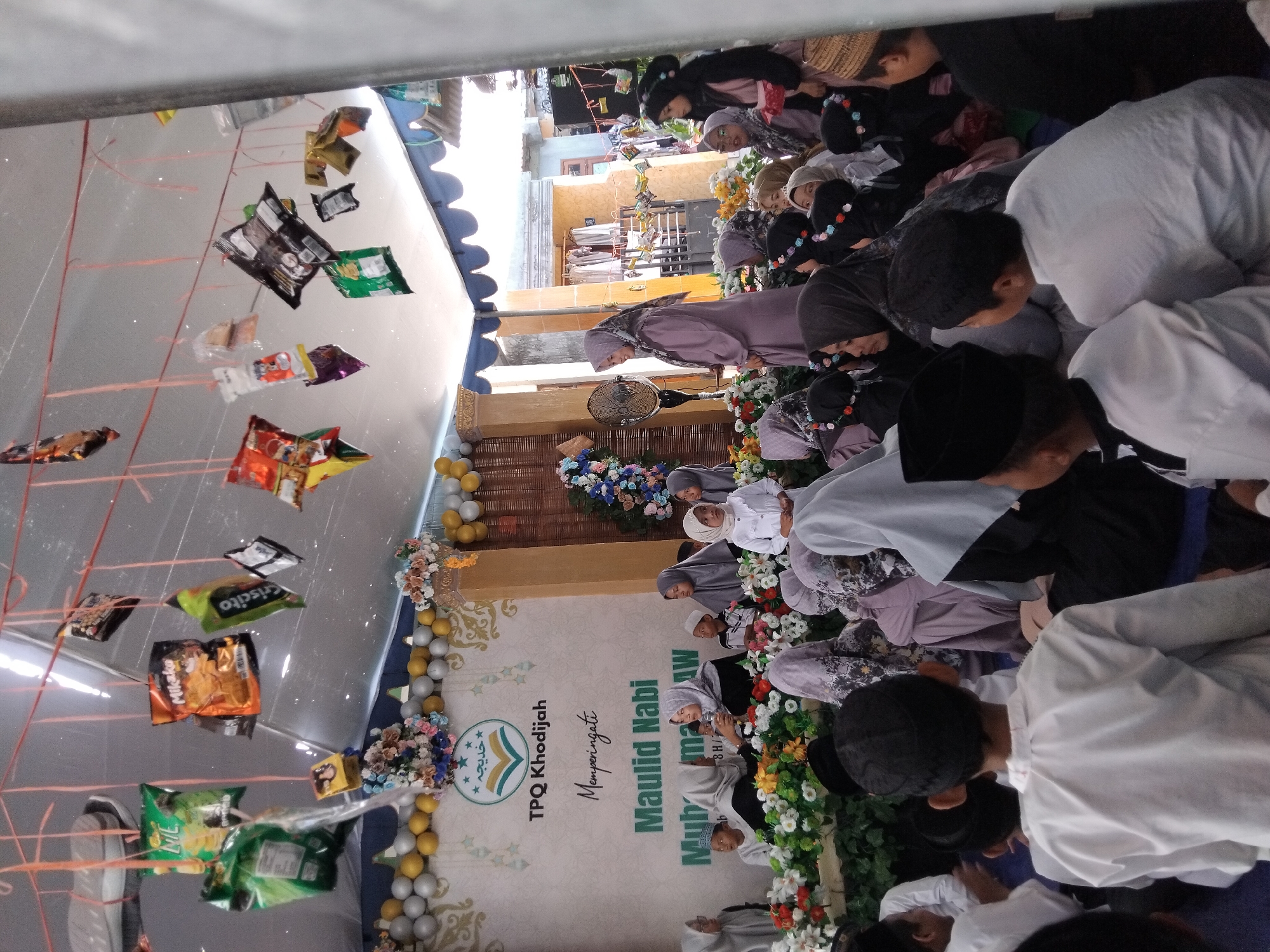 This image captures a scene within an indoor event, likely a religious gathering, held in Kepuh Kiriman, Indonesia. The event is decorated with a banner that reads "TPQ Khodijah Memperingati Maulid Nabi Muhammad SAW." Several children and adults, predominantly women and girls wearing hijabs, are seated on the floor, facing towards a raised platform. A young girl in a white dress and hijab is positioned on the platform, seemingly being addressed or participating in a program. Another woman in a purple hijab stands on the platform, holding a microphone and addressing the attendees. Suspended from the ceiling are various small, individually wrapped food items, possibly snacks, attached to red strings. The background features a stage decorated with flowers and balloons, and the overall ambiance suggests a community celebration or educational event.