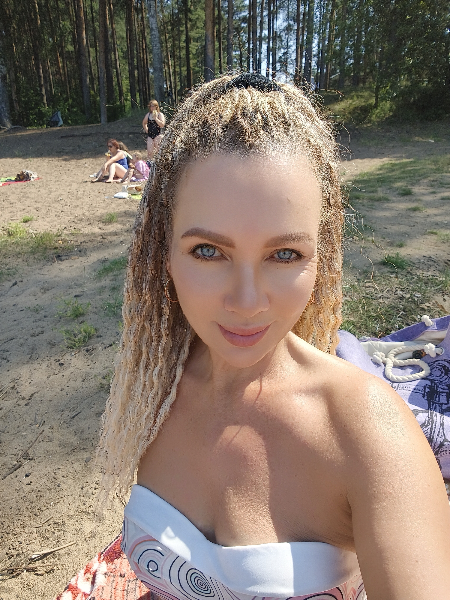 The image is a selfie taken outdoors on a sandy area with trees in the background. The subject, a woman with blonde curly hair and blue eyes, is in the foreground, wearing a white strapless top. She is looking at the camera and smiling. In the background, several other people are visible, some sitting on the sand and others standing. The environment appears to be a beach or a wooded area near water, with sunlight filtering through the trees.