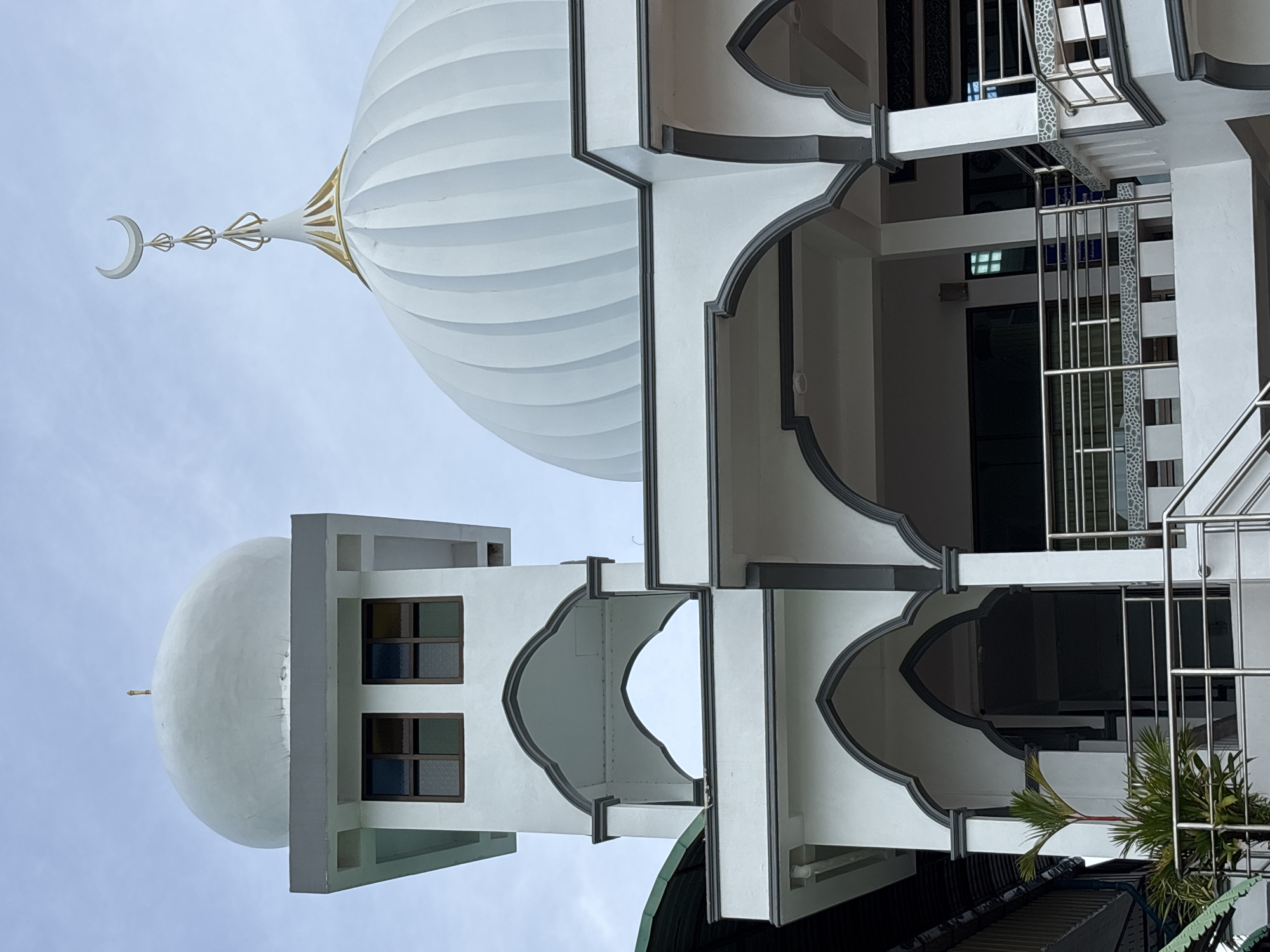 The image shows a low-angle shot of a mosque in Rawai, Thailand, under a cloudy sky. The mosque features white walls with dark gray trim and multiple levels of arched walkways. A tall, square tower with a dome is on the left, and a larger, ribbed dome topped with a crescent moon and finial is on the right. A small palm tree is visible in the lower left corner. The scene is devoid of people.
