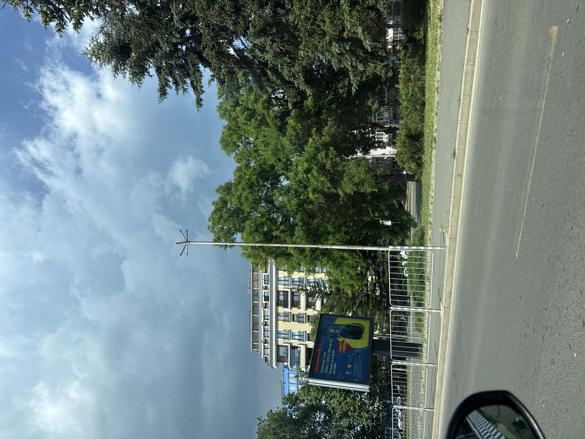 The image depicts a street scene in Burgas, Bulgaria, under a partly cloudy sky. In the foreground, the asphalt road surface is visible, with a yellow lane marking. To the left, the edge of a vehicle's side mirror with a black and white checkered pattern is in view. A concrete curb separates the road from a sidewalk. Behind the sidewalk, a metal railing runs horizontally across the frame. Beyond the railing, a billboard displays an advertisement with text in Cyrillic script and an image of a smartphone. The billboard is situated in front of a building with a light-colored facade and multiple balconies. To the right of the billboard, a tall, slender light pole stands. Further into the background, lush green trees of various sizes dominate the landscape, with a large conifer tree on the far right. The sky above is a mix of light blue patches and dark, heavy clouds, suggesting an approaching storm or recent rain. The overall environment appears to be urban, with elements of greenery and modern architecture.