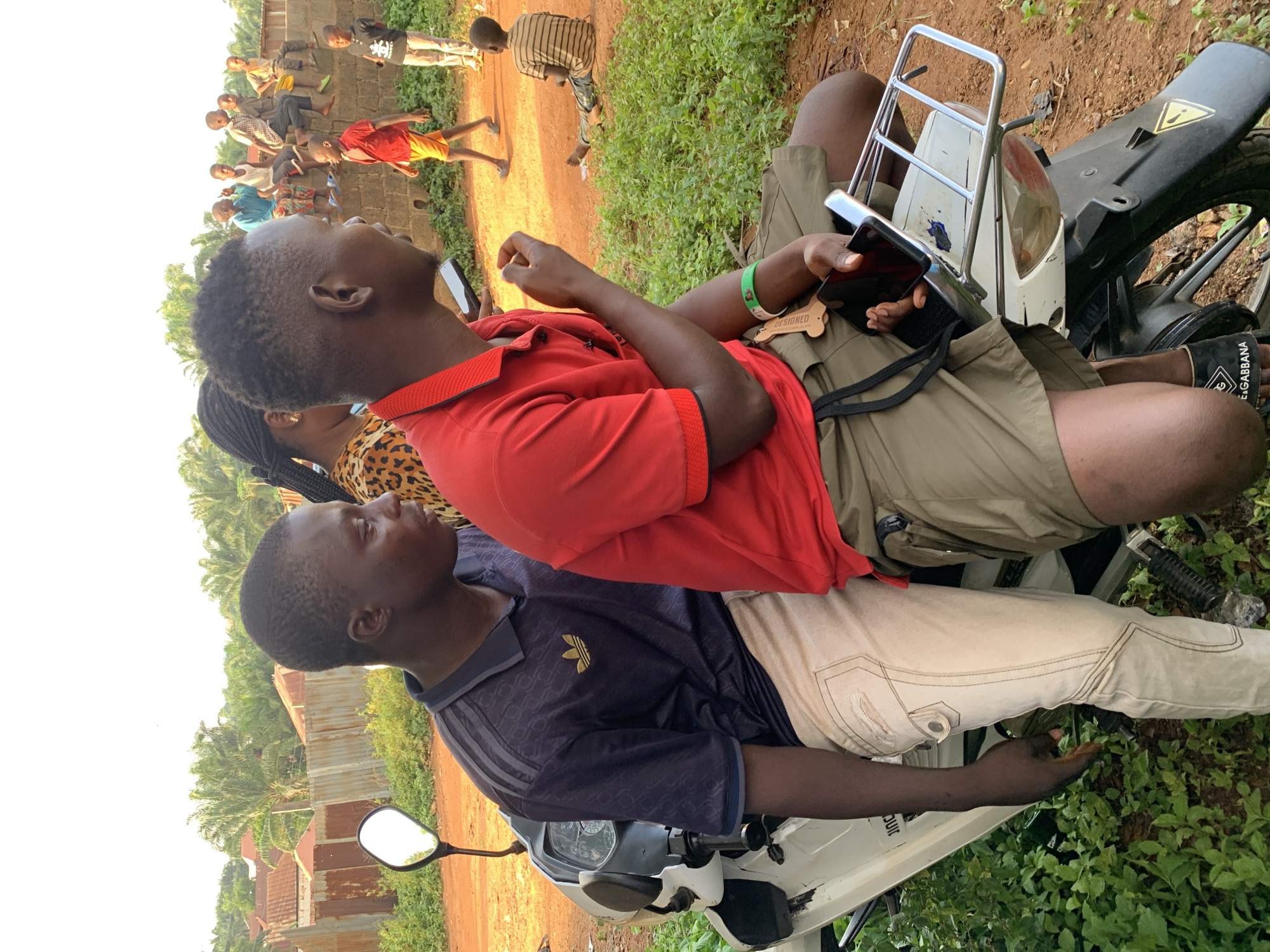In Akwanga, Nigeria, two people are sitting on a white motorcycle. The person in the foreground is wearing a dark blue Adidas polo shirt and light-colored pants. The person behind is wearing a red collared shirt and khaki shorts, and is holding a phone. A group of people are standing on a dirt road in the background. Vegetation surrounds the subjects. Buildings with corrugated metal roofs can be seen in the background.