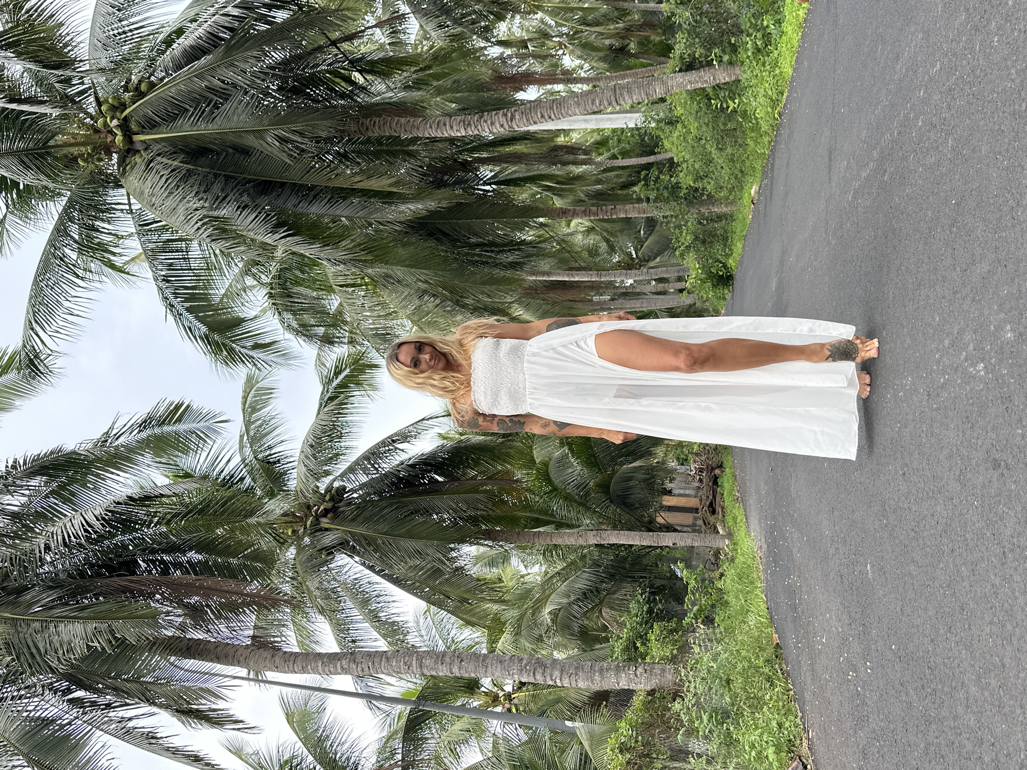 A blonde woman in a white strapless dress with a high slit stands on an asphalt road lined with numerous palm trees. The woman is facing the camera and smiling. Her dress is floor-length and billowy, with a smocked bodice and a sheer section at the thigh. She has tattoos on her arms and her feet are bare, with a tattoo on her left foot. The road appears to be in a rural area, with dense vegetation and some low-lying buildings visible in the background. The sky is overcast, and the overall lighting suggests it is daytime. The scene is consistent with the landscape found in Mui Ne, Vietnam, known for its coastal scenery and extensive coconut palm plantations.