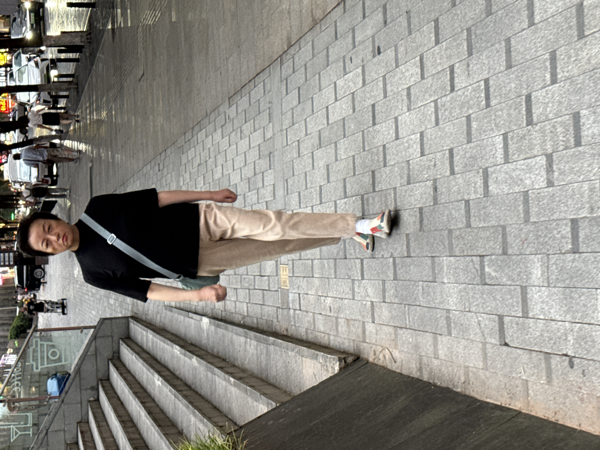 A full-shot, eye-level, medium-distance photograph shows a middle-aged East Asian woman walking on a cobblestone sidewalk in Yuzhong District, China. The woman is wearing a black short-sleeved shirt, tan pants, and multi-colored sneakers. A light blue crossbody bag is slung over her right shoulder. Her hair is short and dark, and her facial expression is neutral, with her lips slightly pursed. She is walking towards the viewer, and the background features a blurred street with vehicles and other pedestrians, as well as a set of stone stairs to the left of the frame. The lighting suggests it is either early morning or late afternoon, with dappled sunlight creating shadows on the sidewalk. The overall scene depicts an ordinary urban moment.