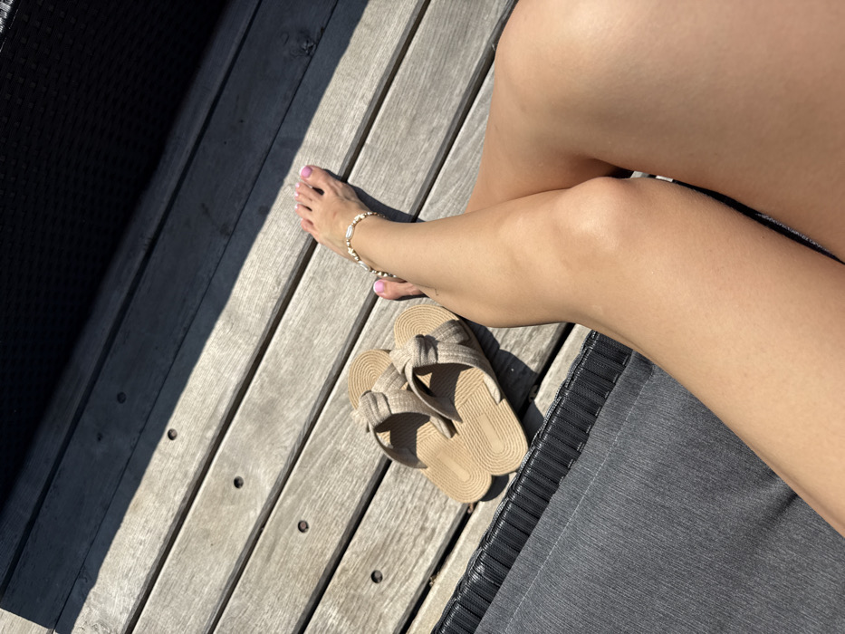 The image captures a close-up view of the lower legs and feet of an individual, resting on a dark grey outdoor lounge chair in Hatne, Ukraine. The individual's legs are crossed at the knees, exhibiting tanned skin. The visible right foot has toenails painted pink, and an ankle bracelet adorned with white and gold-toned beads is worn on the right ankle. Adjacent to the individual's feet, on a light-colored wooden deck, a pair of beige slide sandals with woven-patterned straps are placed. The wooden deck consists of parallel planks, and strong sunlight casts defined shadows, including a large dark shadow in the upper right portion of the frame.