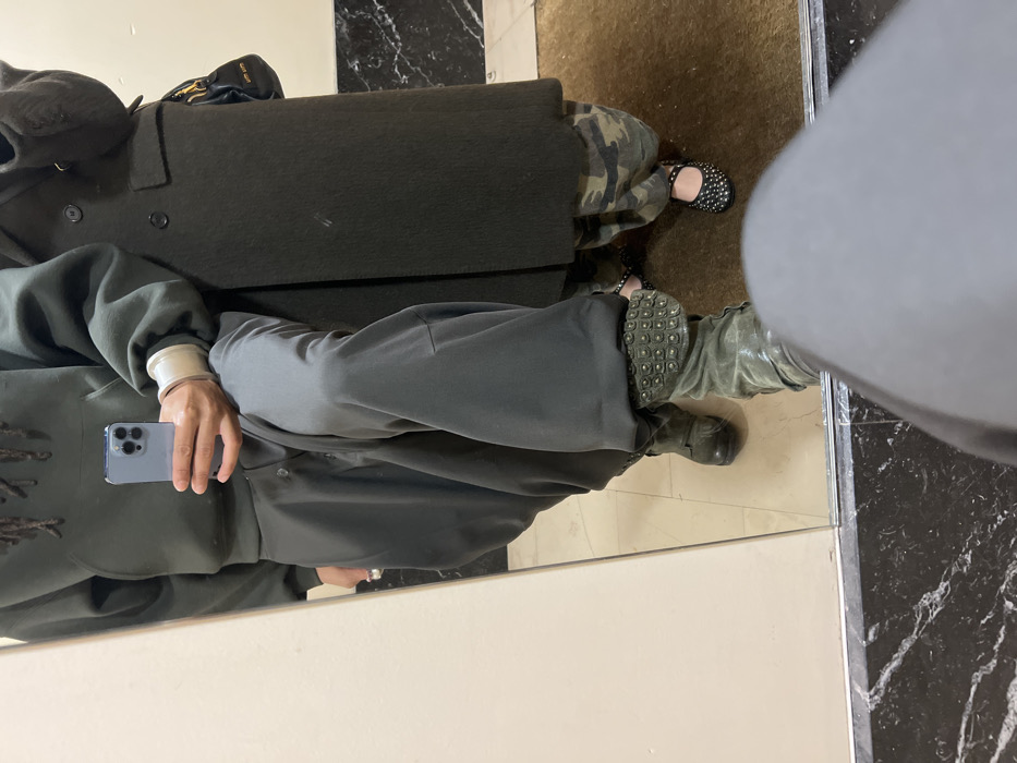 The image displays a mirror selfie of two individuals, partially visible from approximately the chest down, in an indoor setting in Paris, France.
On the left side of the reflection, one individual holds a light blue smartphone with their right hand, positioned to capture the image. This person wears a dark green hooded sweatshirt, dark gray wide-leg pants, and olive-green boots. The right boot features a prominent studded cuff. A white wristband is visible on the individual's left wrist, and a silver-toned ring is on the left ring finger. Above the hoodie, a portion of dreadlocks is visible.
To the right, a second individual stands, wearing a dark, long coat secured with two visible buttons, camouflage-patterned pants, and black Mary Jane-style flats adorned with studs. A black handbag with a yellow detail is slung over their right shoulder.
The floor in the foreground consists of a dark, polished marble-like surface with prominent white veining, transitioning into a plain brown mat or carpet. The mirror itself reflects an off-white wall section on the left and a darker, less distinct surface on the right.