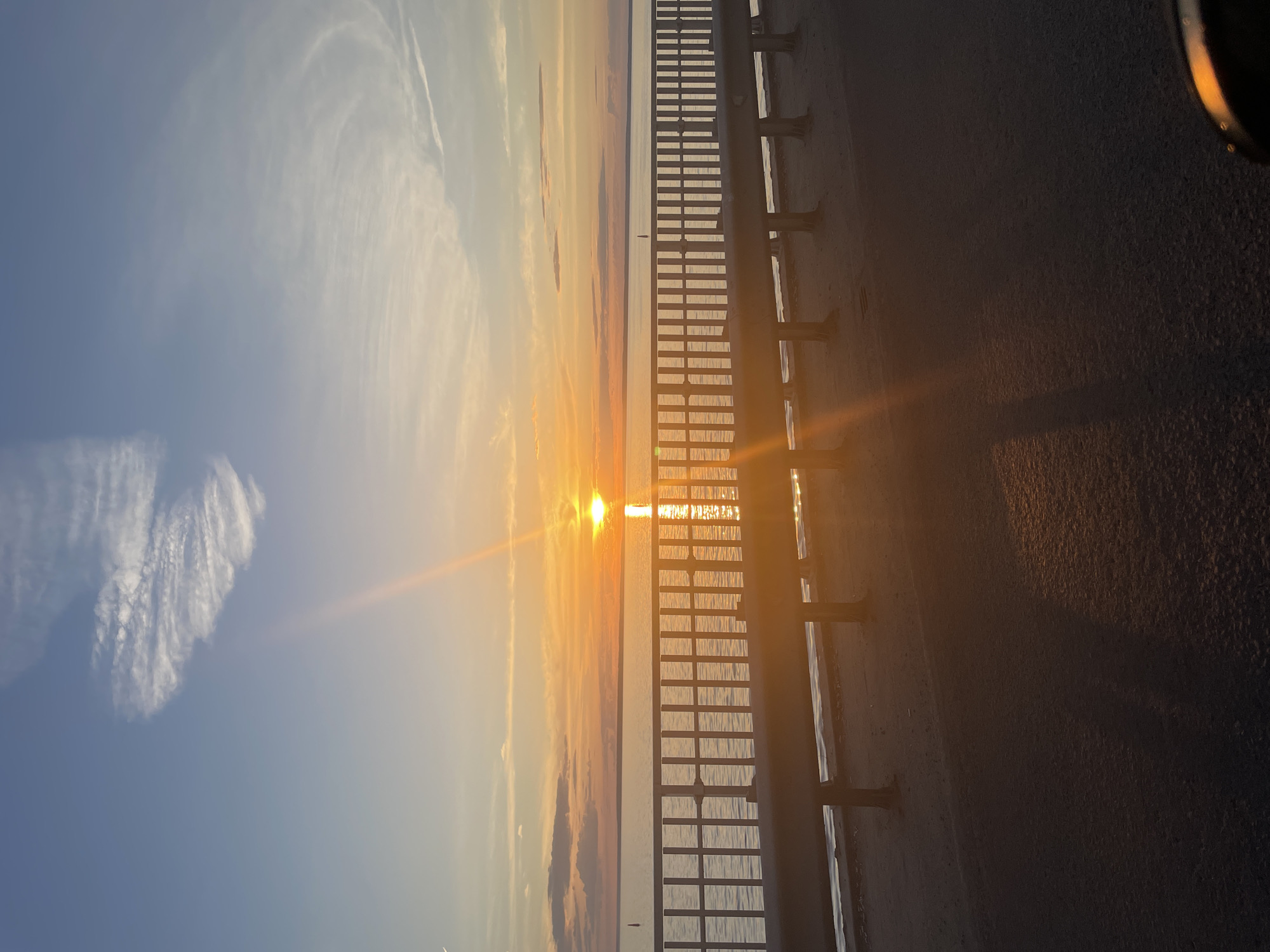 The image shows a sunset over a body of water, as viewed from a bridge or overpass. The sun is low on the horizon, casting a bright orange glow across the sky and reflecting on the water. The sky is a gradient of blue and orange, with wispy clouds. A metal railing runs horizontally across the foreground, separating the viewer from the water. Below the railing, there is a sandy area or beach. The road surface is visible in the very bottom of the frame. The scene is peaceful and serene, with the warm colors of the sunset creating a tranquil atmosphere. There are no people or specific landmarks visible that would help to identify the location within the city of Unknown, Unknown.