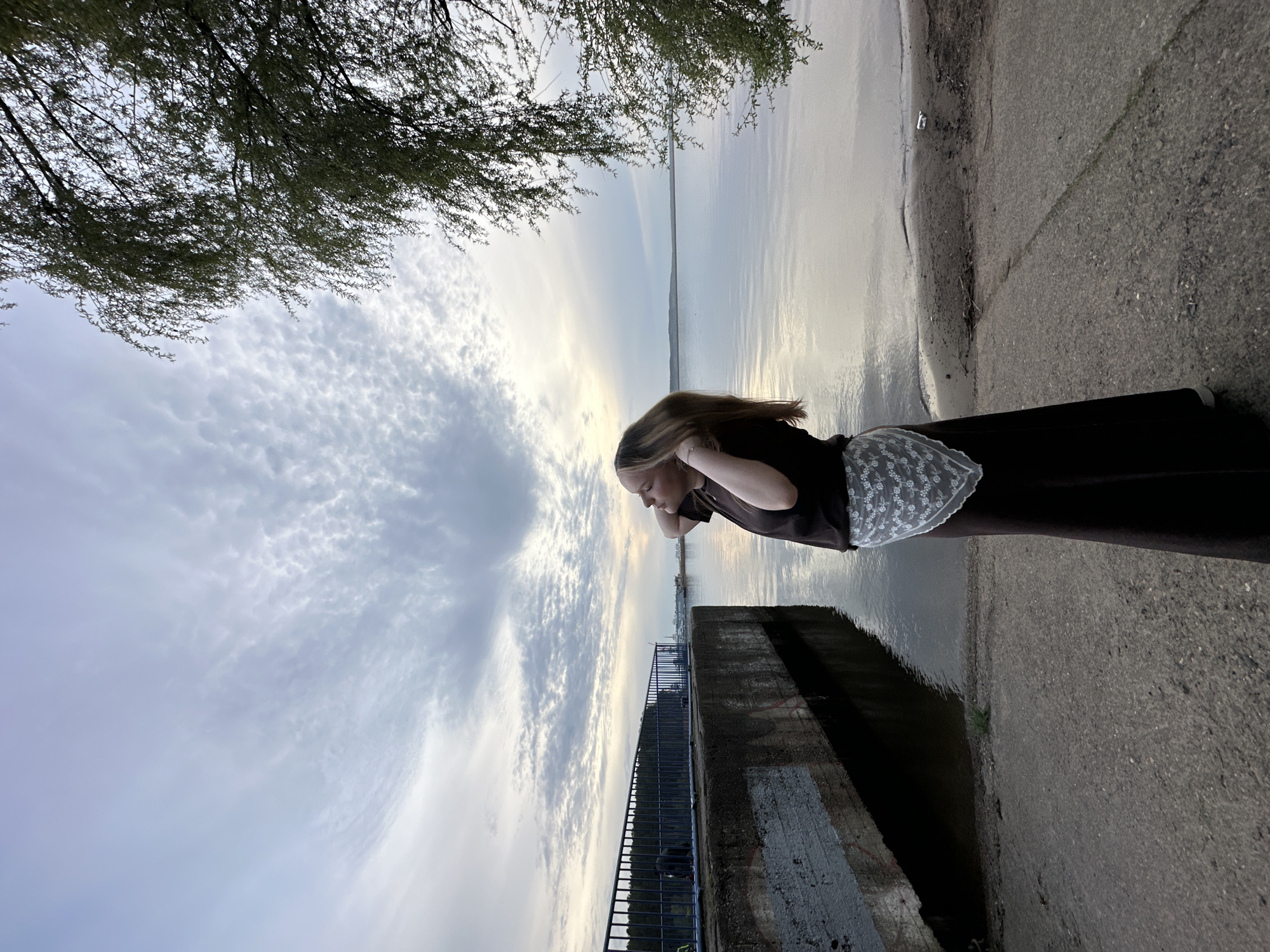 A young woman with long blonde hair is standing by a body of water, possibly a lake or reservoir, in the city of Chaikovsky, Russia. She is wearing a dark brown or black dress with a white lace detail at the back. Her hands are near her head. She is looking down towards the water. A concrete structure with a metal railing is visible near the water. The sky is cloudy with a mix of gray and white. Branches of a tree are visible in the upper left corner of the image. The photograph is taken at ground level and is rotated. The light suggests that the picture was taken either during sunrise or sunset.