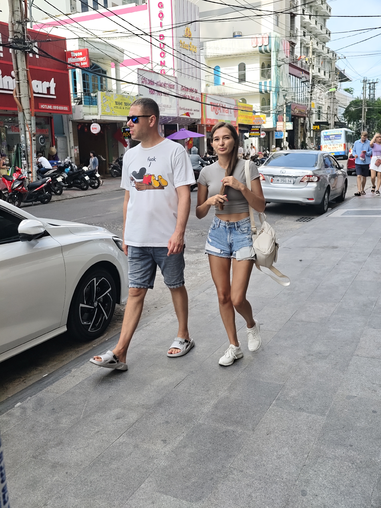 Two individuals, a man and a woman, walk along a city sidewalk in Nha Trang Ward, Vietnam. The man, wearing a white t-shirt with a graphic print and denim shorts, walks with his left leg forward. The woman, in a grey cropped t-shirt and distressed denim shorts, walks slightly behind and to his right, carrying a beige backpack. Both are wearing casual footwear. The background reveals a street scene with buildings, parked motorcycles, and a silver sedan. The pavement is made of large grey tiles. Overhead, a dense network of electrical wires crisscrosses the sky.