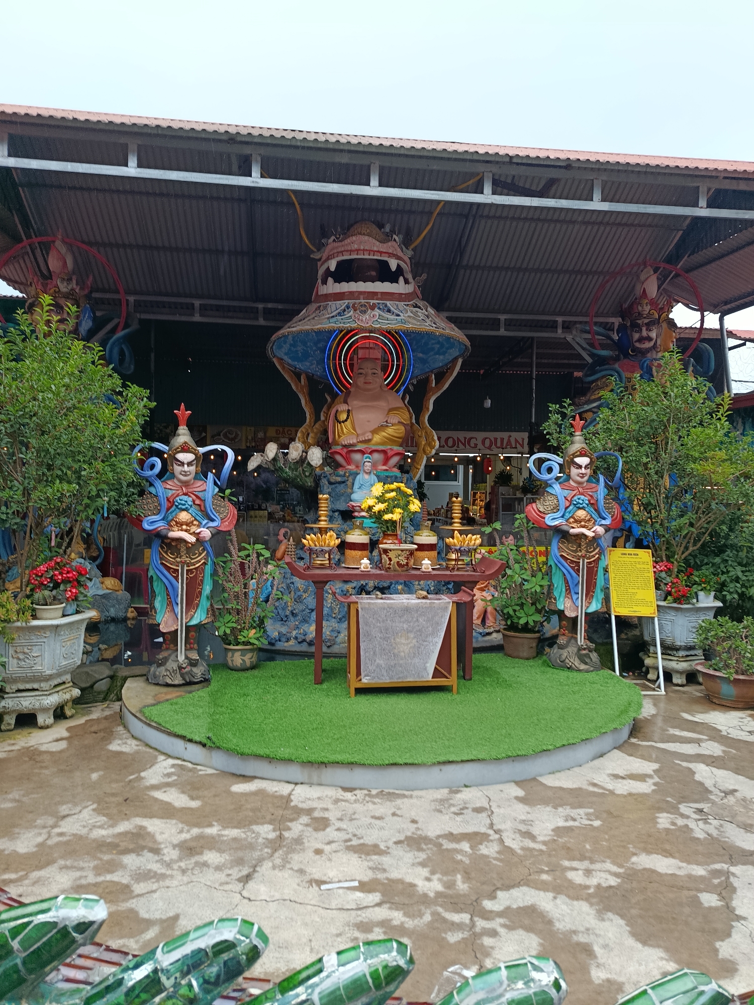 This image shows an outdoor shrine in Xuân Thành, Vietnam, likely a Buddhist temple or a similar religious site. The central focus is a large Buddha statue seated under a decorative, conical canopy adorned with colorful neon lights, including a circular halo effect. Flanking the Buddha are two imposing statues of warriors or guardians, each holding a sword and wearing ornate armor and helmets.
In front of the Buddha is a low, red altar table covered with offerings, including yellow flowers, incense sticks, and other decorative items. A smaller statue of a deity or monk is positioned behind the altar table. The shrine is set on a raised, circular platform covered with artificial green turf.
To the left and right of the main structure are lush green trees and potted plants. Several other statues and decorative elements are visible in the background, including what appears to be a dragon motif on the canopy and large, imposing figures on either side of the roofline. A yellow sign with Vietnamese text is prominently displayed to the right of the right guardian statue.
The ground surrounding the turf platform is paved with concrete that appears wet and cracked, suggesting it may have recently rained or been washed. In the foreground, partially visible, are several decorative, green, segmented objects, possibly part of a larger installation or railing. The overall atmosphere is one of solemnity and reverence, typical of a religious sanctuary.