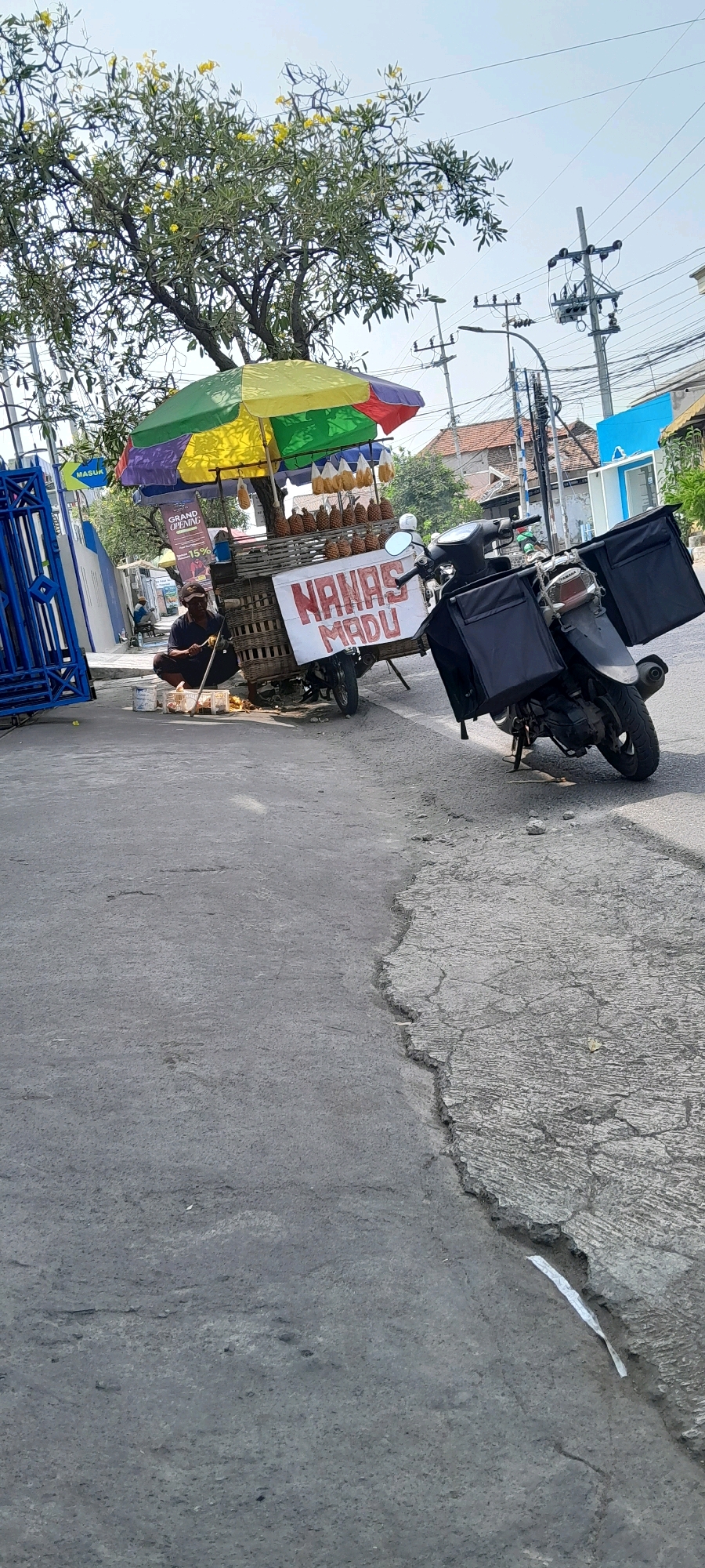 The image depicts a street scene in Pasuruan, Indonesia. A vendor is operating a pineapple cart under a multi-colored umbrella, displaying ripe pineapples and some hanging fruit. The cart is laden with pineapples in woven baskets, with a prominent sign in red text reading "NANAS MADU". A motorcycle with two large black storage boxes is parked next to the cart. A person, presumably the vendor, is seated near the cart, actively engaged in preparing fruit, with a small collection of containers and utensils on the ground. In the background, there is a tree with yellow flowers, electrical poles, and buildings. The street itself is paved and shows signs of wear and tear.