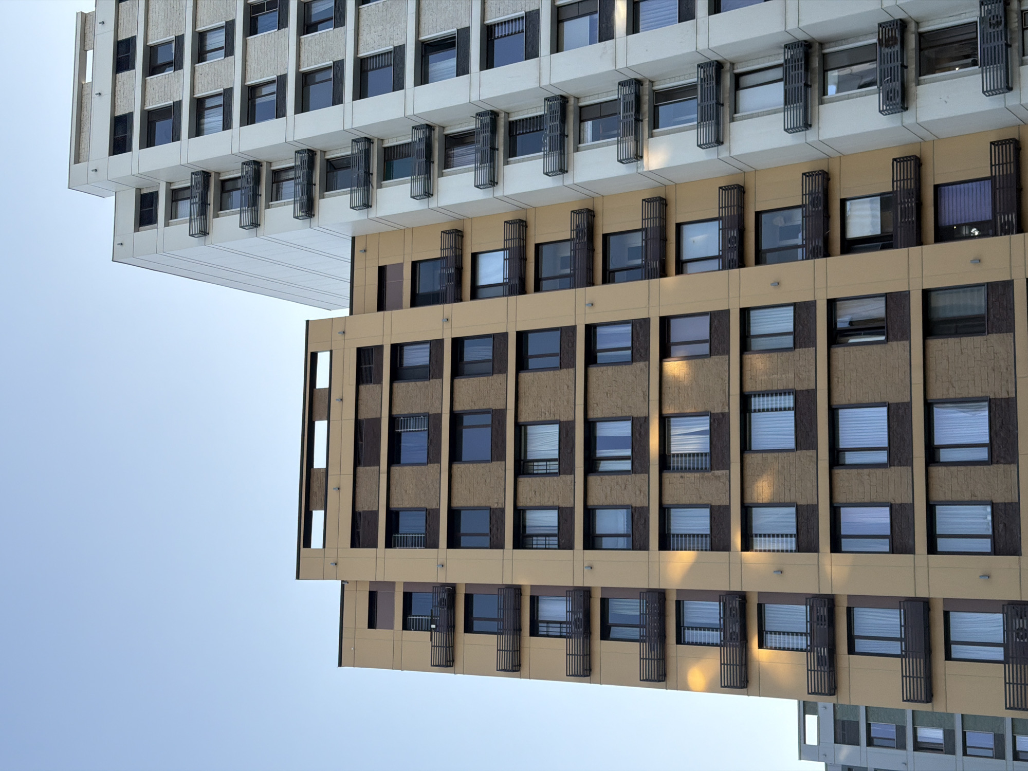 This image depicts the exterior of modern apartment buildings, likely in Almaty, Kazakhstan, under a clear, pale blue sky. The primary focus is on two adjacent buildings: a taller, light-colored building on the right and a slightly shorter, tan-colored building on the left.
The light-colored building on the right features multiple floors with a repeating pattern of windows and small balconies with dark railings. The facade is a mix of smooth, off-white panels and textured stone-like elements. The windows are rectangular and appear to be fitted with blinds or curtains.
The tan-colored building on the left has a different architectural style. Its facade is predominantly tan with dark brown accents and bands running horizontally between floors. This building also has many windows, and some floors feature balconies with dark railings, similar to the adjacent building. The windows on this building appear to be more varied in their coverings, with some showing sheer white curtains and others appearing darker or reflective.
In the background, to the far left, parts of another building, possibly also an apartment complex, are visible. This building has a lighter grey facade and a more industrial appearance, with rows of windows that seem to be recessed.
The lighting suggests it is daytime, possibly late afternoon, as there are some shadows and highlights visible on the facades, particularly on the tan building where sunlight creates streaks of light. There are no people or vehicles visible in the image. The scene is a static architectural landscape, devoid of any human activity.