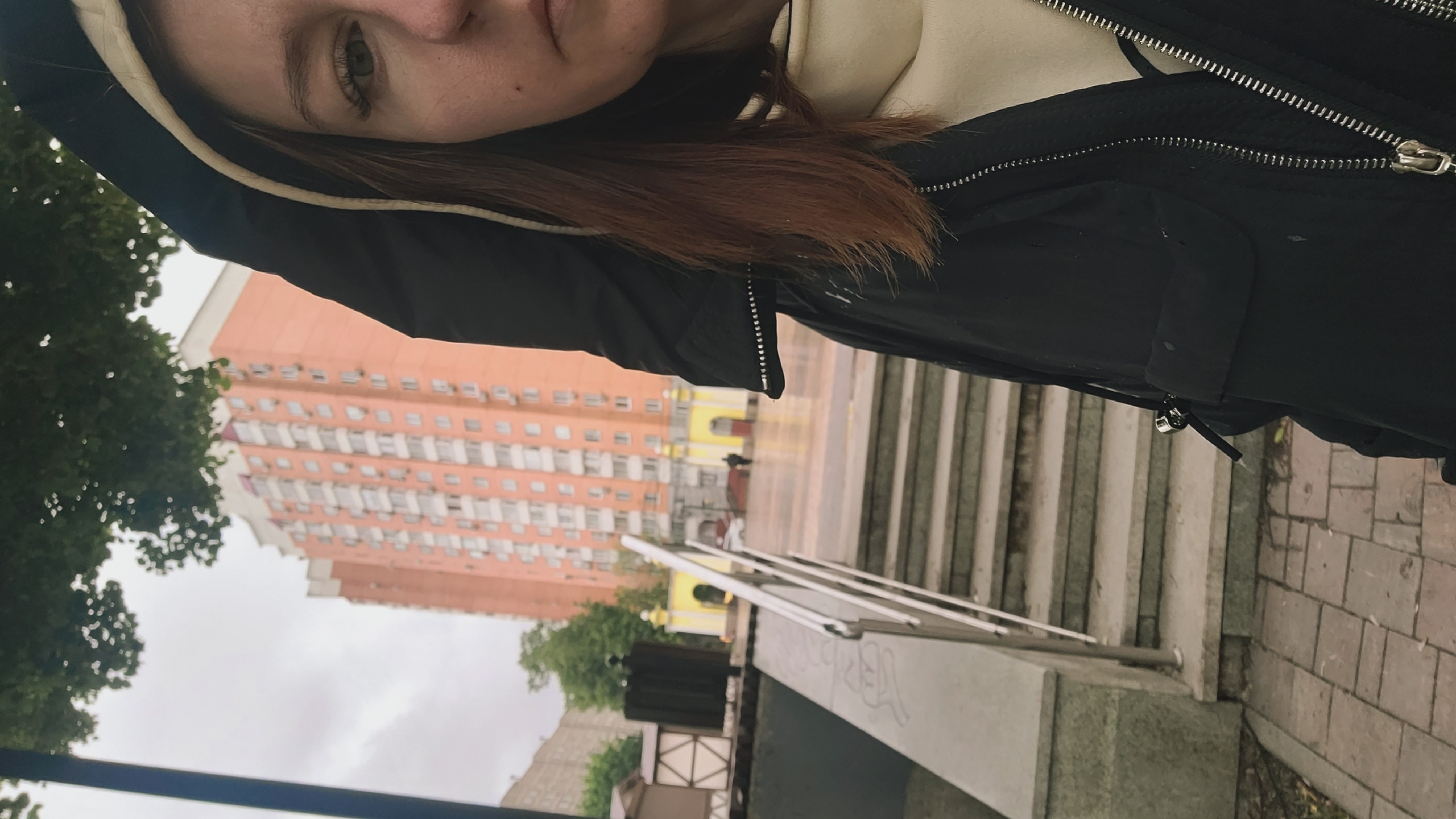 The image captures a woman standing outside on a cloudy day, seemingly in an urban environment in the city of Unknown, Unknown. She is wearing a black jacket with a hood and a beige hoodie underneath. Her hair is reddish-brown and partially obscured by the hood. In the background, there is a tall, pinkish-orange building with numerous windows. Also visible are a set of stairs leading up to a building with a yellow archway, a metal railing, a paved sidewalk, and patches of green trees. The overall impression is that the photo was taken during a walk in a cityscape, possibly after or during light rain, considering the slightly wet surfaces.