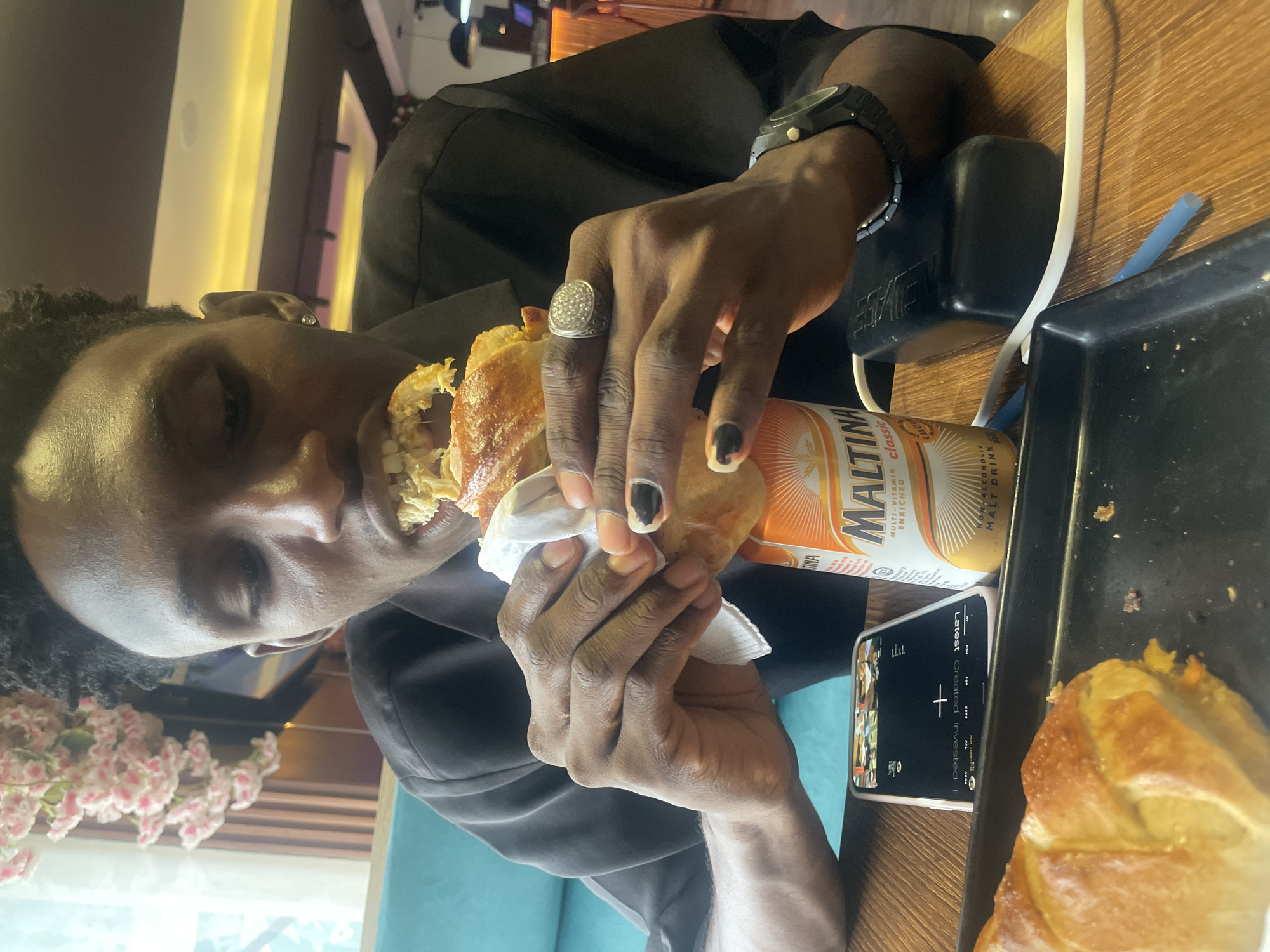 A man is eating a sandwich, holding it in both hands, with a can of Maltina malt drink next to his phone on a wooden table. The man is wearing a black shirt and a large watch, and a ring. The table is set with a blue placemat. Flowers can be seen in the background.
