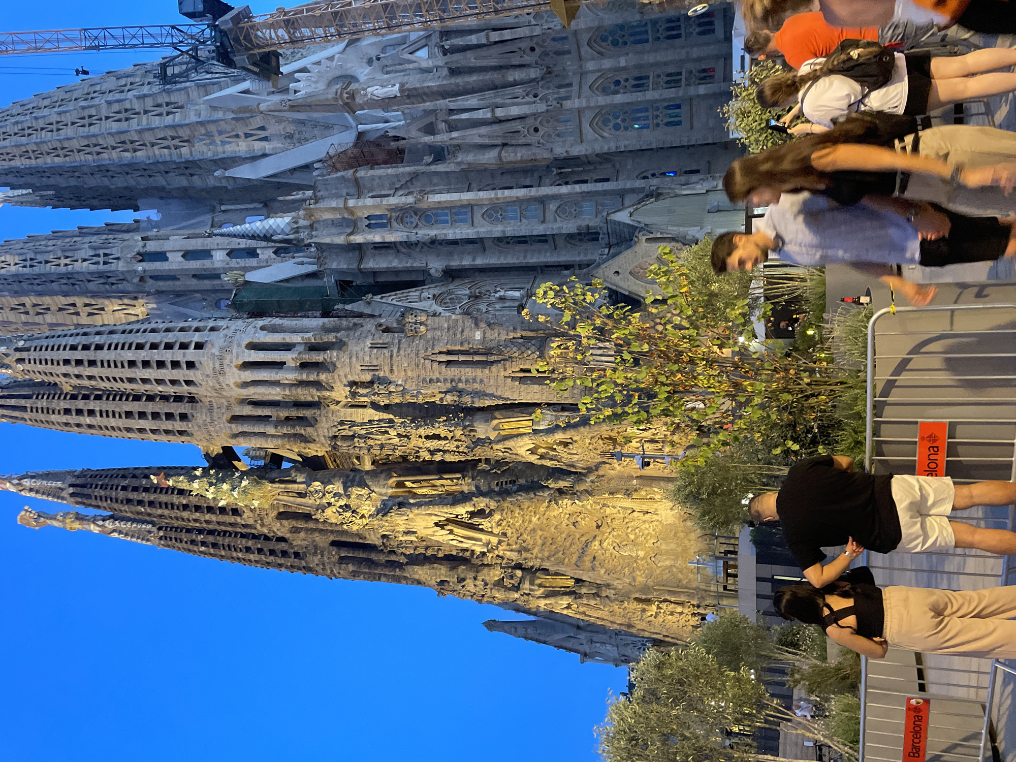 This image captures a frontal view of the Sagrada Familia basilica in Barcelona, Spain, at dusk. The sky is a deep blue, and the upper portions of the basilica's towers are illuminated with a warm yellow light. The facade is intricately detailed with Gothic and Art Nouveau architectural elements. In the foreground, several people are visible. On the left, a couple stands facing away from the camera, with the woman wearing a black top and beige pants, and the man in a black t-shirt and white shorts. To their right, a man in a light blue shirt and black shorts walks past, followed by a woman in a black tank top and beige pants, who is taking a photo with her phone. Further to the right, another person in an orange shirt is partially visible. A metal barrier with red signs reading "Barcelona" runs across the lower part of the image. Trees and decorative plants are interspersed between the people and the basilica. A construction crane is visible on the right side of the basilica, indicating ongoing work.