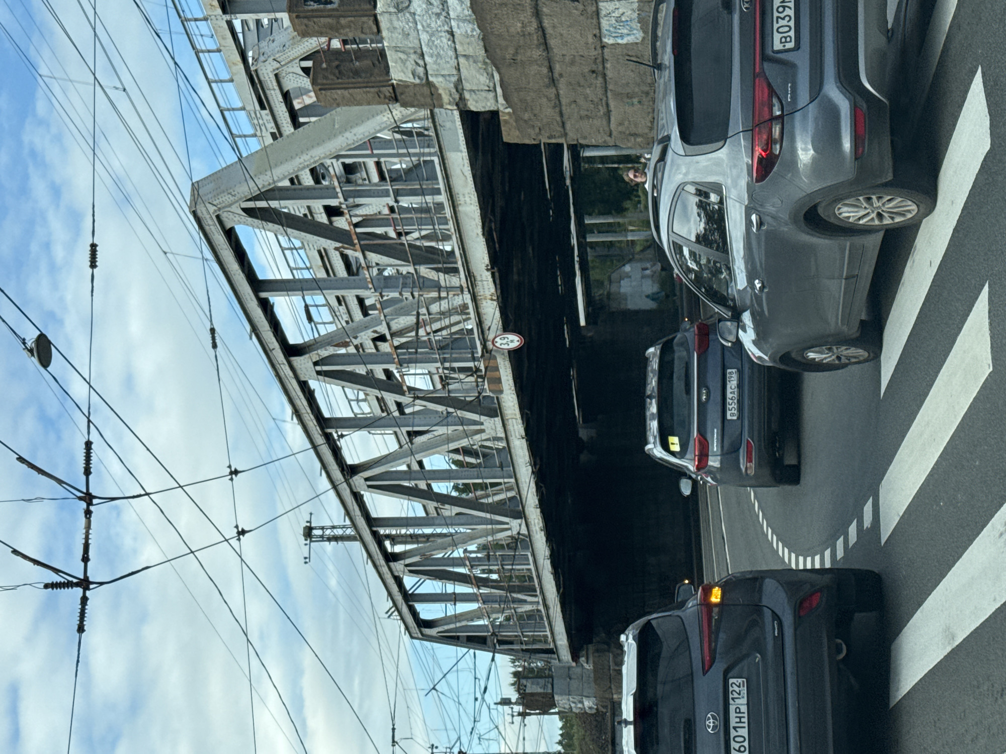 The image shows a view from a car driving on a road under a large, industrial railway bridge. The bridge is made of steel trusses and spans across the road. There are multiple sets of overhead wires for what appears to be an electric railway. Several cars are visible on the road, including a dark gray Toyota SUV in the foreground on the left and a gray Kia SUV in the middle. The road has white dashed lines indicating lanes and a pedestrian crossing. In the background, beyond the bridge, trees are visible. There are also some people standing near the side of the road. The sky is partly cloudy. The license plate of the Toyota SUV reads "601HP 122" and the license plate of the Kia SUV reads "B556AC 198". The license plate on the grey Kia Rio in the right is B039M. A circular sign with "3.9 M" is visible on the underside of the bridge, indicating a height restriction. The scene is likely in Saint Petersburg, Russia, given the context of the image and typical road infrastructure found there.