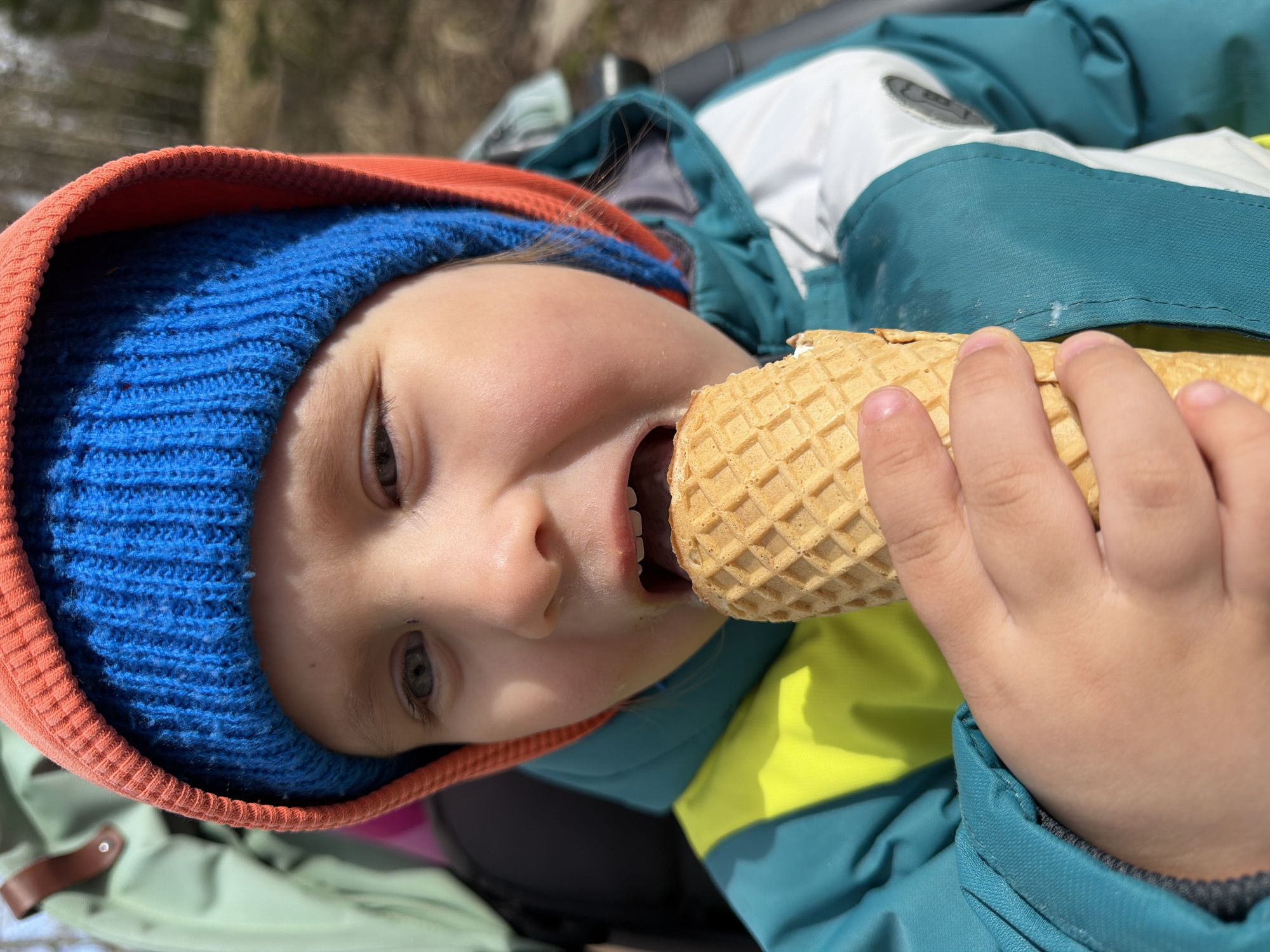 A young child with light skin and gray-green eyes, wearing a blue knitted hat with an orange rim and a teal, lime green, and white jacket, is eating an ice cream cone with a waffle cone in an outdoor setting. The child is tilted slightly to the side, with the cone nearly touching their lips. The background includes foliage, and it is daytime. The location is not explicitly provided and cannot be determined from the image alone.
