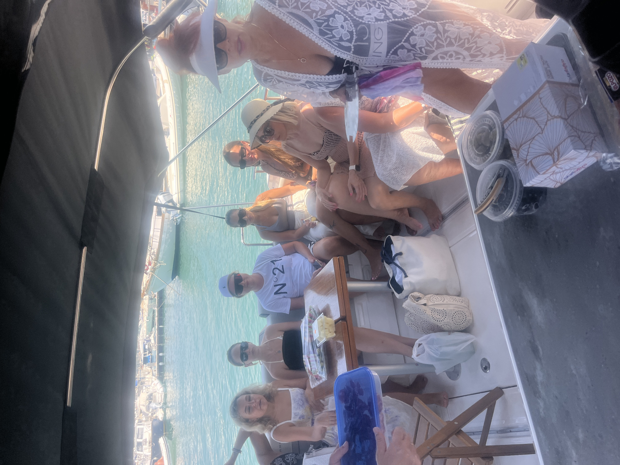 A group of six people are on a boat in the sea. The people are mostly women, dressed in summer attire. Some are seated at a wooden table on the boat, while others are standing. There are various items on the table, including food containers, a wine opener, and bags. The background shows a harbor with several boats and buildings. The water is a clear turquoise color, and the sky is bright and sunny. The overall scene suggests a leisurely outing on a boat. The city of Polis Chrysochous, Cyprus is known for its picturesque harbor and clear waters, making it a plausible location for such an activity.