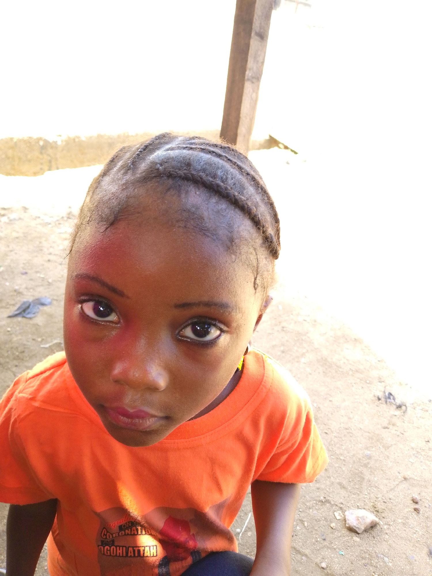 A young girl with braided hair is looking up at the camera. She has dark eyes and is wearing an orange t-shirt with a graphic on it. The background appears to be an outdoor area, possibly in the city of Lokoja, Nigeria, with sandy ground and a wooden post visible.