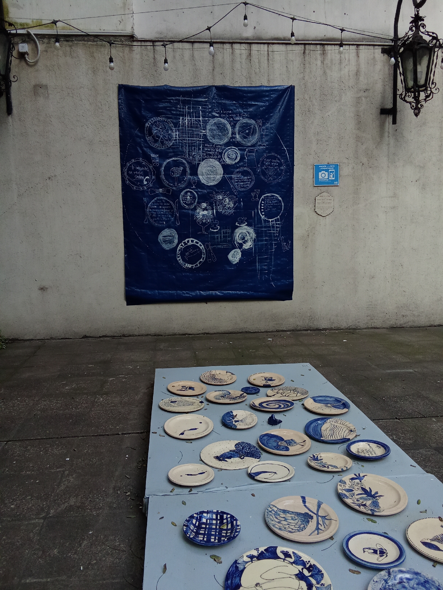 The image depicts an outdoor exhibition of ceramic plates, displayed on a light blue platform against a textured off-white wall. The wall is adorned with a large blue tarp featuring white line drawings of plates and other objects, resembling a blueprint. String lights hang overhead. A decorative black lantern is mounted on the right side of the wall, above a small blue and white sign with a camera icon. Scattered on the ground are fallen leaves. The scene is likely in an urban setting in Buenos Aires, Argentina, possibly a courtyard or a public art space. There are no people visible in the image.