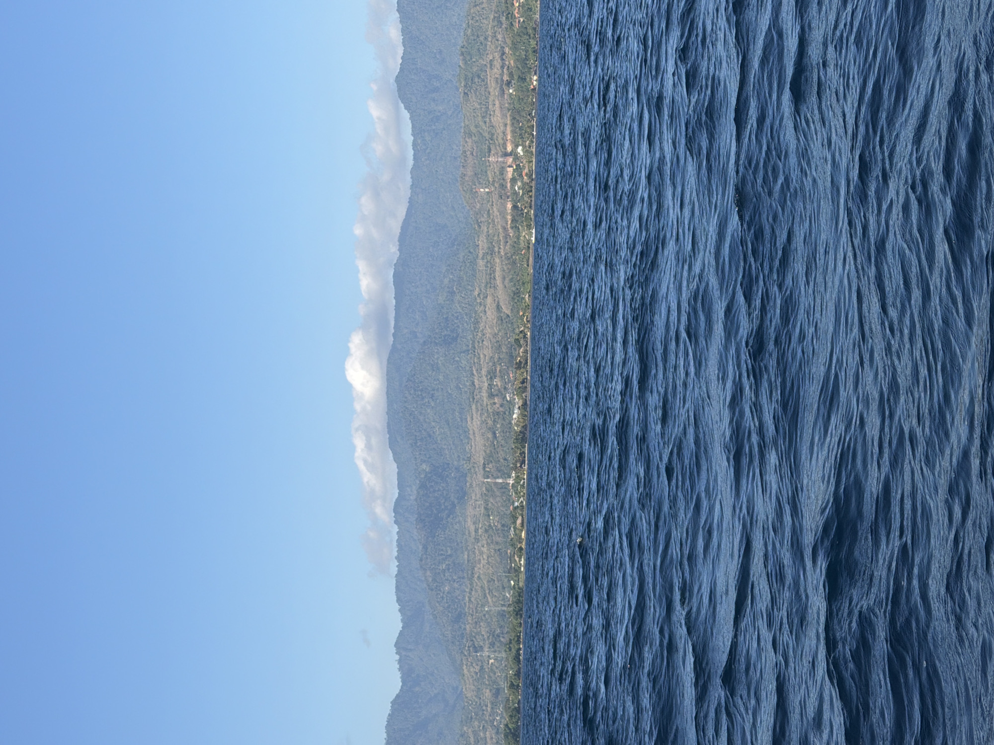 The image depicts a view of the sea and the coastline of Seririt, Indonesia. The foreground is dominated by deep blue ocean water with visible ripples and waves. In the midground, a low-lying coastline is visible, characterized by lush green vegetation and scattered buildings. Several tall transmission towers are discernible along the coast. Behind the coastline, a range of verdant, rolling hills and mountains rises, their slopes covered in dense foliage. The sky above is a clear, bright blue, with a few scattered white clouds lining the horizon above the mountains. The scene appears to be a daytime view, likely taken from a boat on the water.