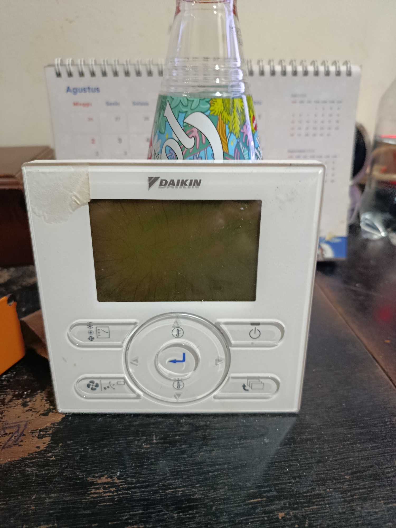 The image shows a white Daikin air conditioning thermostat on a dark wooden surface. The thermostat has a digital display and several buttons, including a power button, temperature control buttons, and fan speed settings. In the background, a calendar with "Agustus" visible and a decorative plastic bottle are present. The scene appears to be indoors, possibly in an office or residential setting. No people are visible in the image. The overall impression is one of a functional but slightly worn electronic device in a domestic or office environment. The context of Central Jakarta, Indonesia, is not visually apparent from the image itself.
