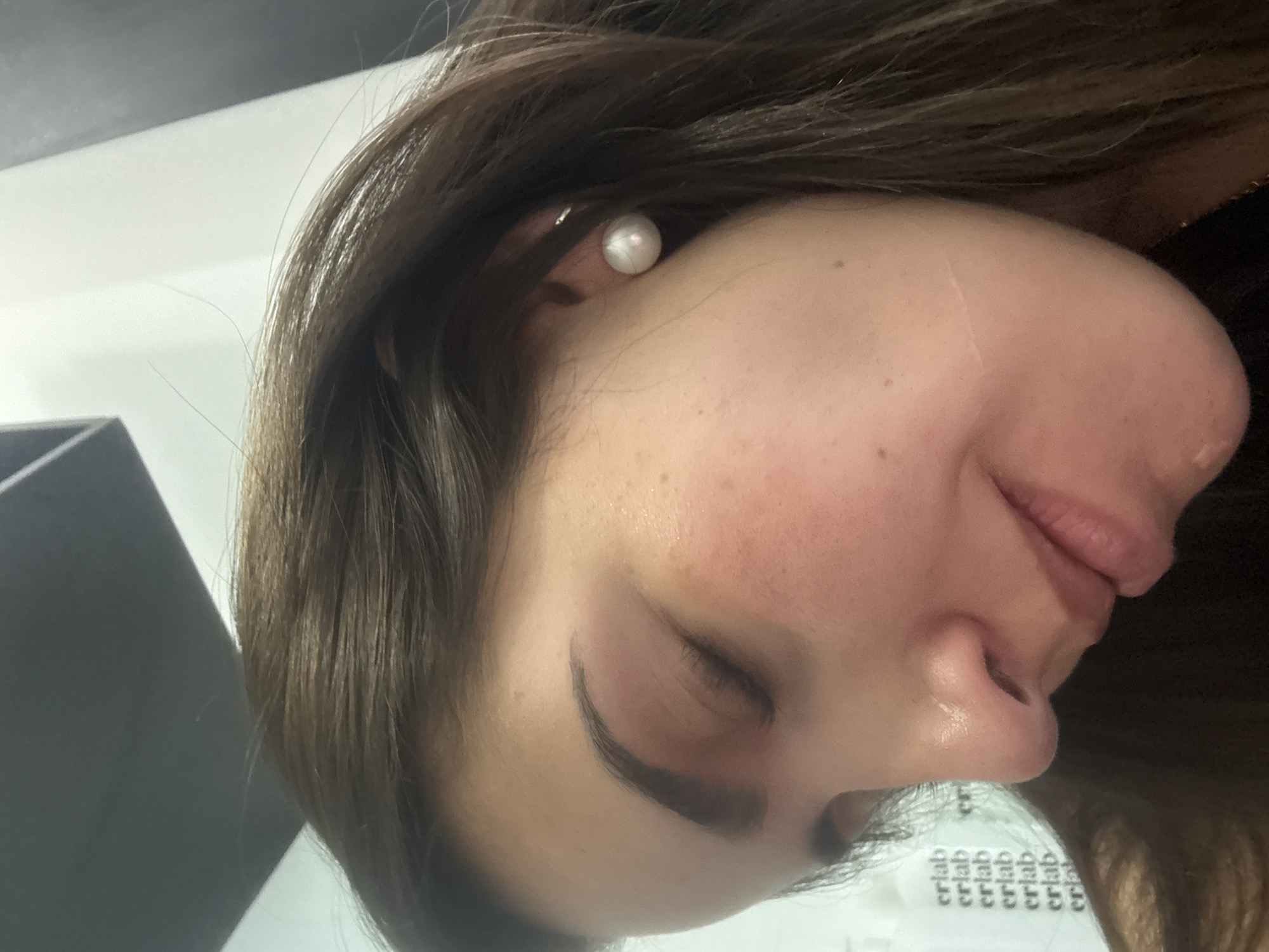 A close-up, rotated view of a person's face. The person has brown hair, fair skin, and is wearing a pearl earring. Their eyes are closed. Part of a black object is visible in the left of the frame. The bottom of the image contains some blurred text. The location in San Bernardino Tlaxcalancingo, Mexico is not discernible from the image.