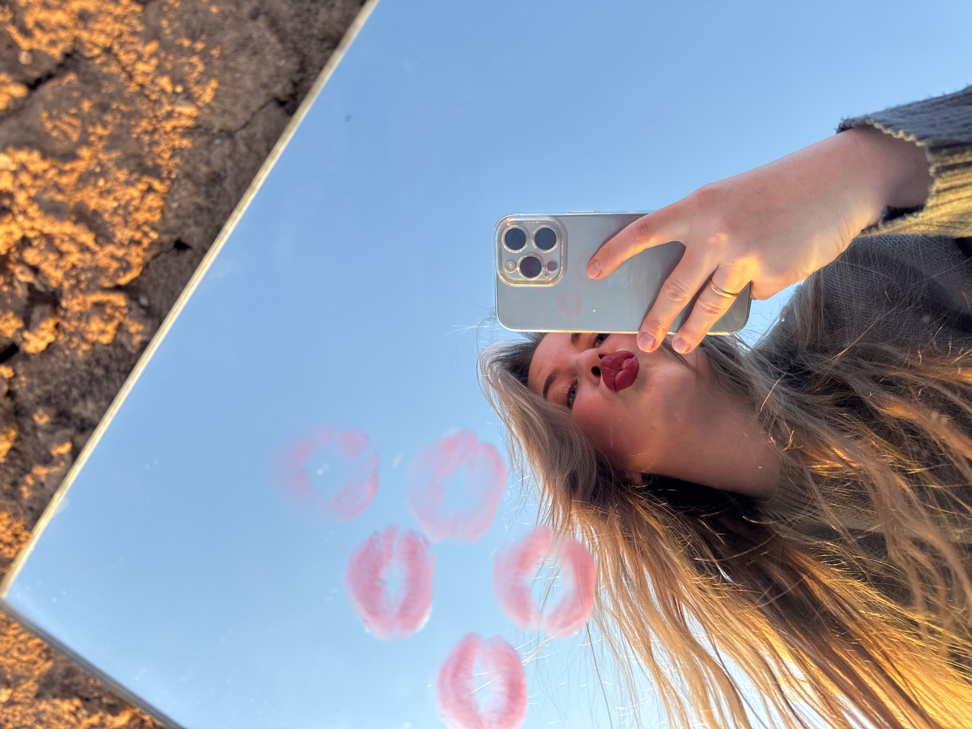 A woman is taking a selfie in a mirror, which is placed on rough, cracked terrain. The mirror reflects a clear blue sky and contains several pink lipstick kiss prints. The woman's face is partially visible in the top right corner of the mirror, showing her lips, nose, and a portion of her hair. She is holding a smartphone with three rear cameras to take the picture. A thin gold ring is on her left hand. The location is possibly in Ольховка, Russia.