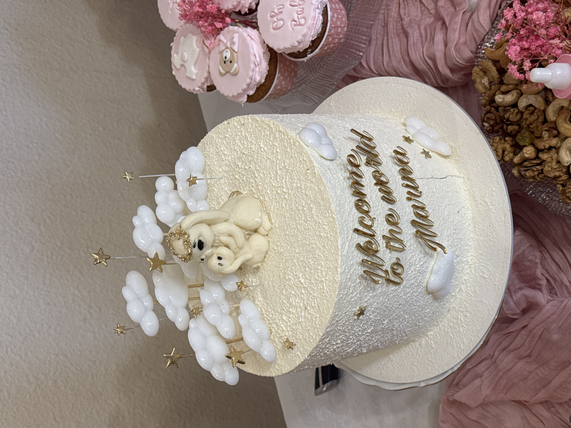 This is a close-up, eye-level, medium shot of a cream-colored, textured cake decorated with edible clouds and gold stars, set against a blurred background. The cake is adorned with a fondant decoration of two white rabbits, one larger than the other, with the larger rabbit wearing a gold crown. Gold lettering on the side of the cake reads, "Welcome to the world Marina". To the right of the cake, several pink-frosted cupcakes are visible, some decorated with teddy bears and others with the text "Oh Baby". A tray of mixed nuts and pink flowers is in the lower right corner of the frame. The scene appears to be an indoor setting, possibly a party or celebration. There is no identifiable information suggesting the location is in Yerevan, Armenia.