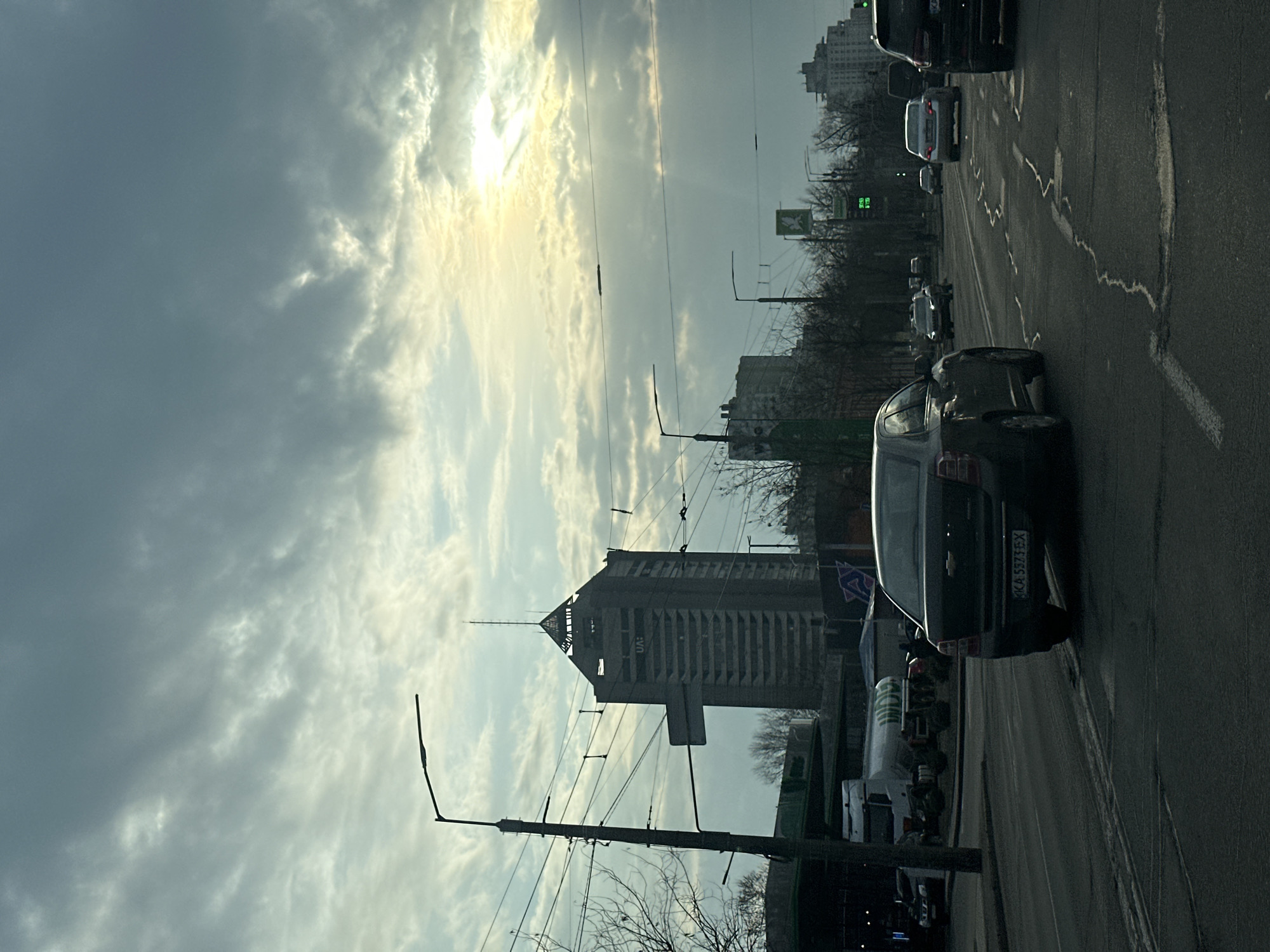 The image is taken from inside a car traveling on a city street, likely in Kyiv, Ukraine, based on the license plate of the car in front (KA 5373 EX), buildings, and other details. The sky is overcast with sunlight breaking through the clouds. There is traffic on the road including several cars and a truck. In the background, there's a distinctive tall building that appears to be the Ukrainian Institute of Scientific and Technical Expertise and Information, sometimes referred to as "UFO" building due to its unique shape. Power lines and street lamps are visible along the roadside. Some trees are visible as well with bare branches, suggesting a colder season.