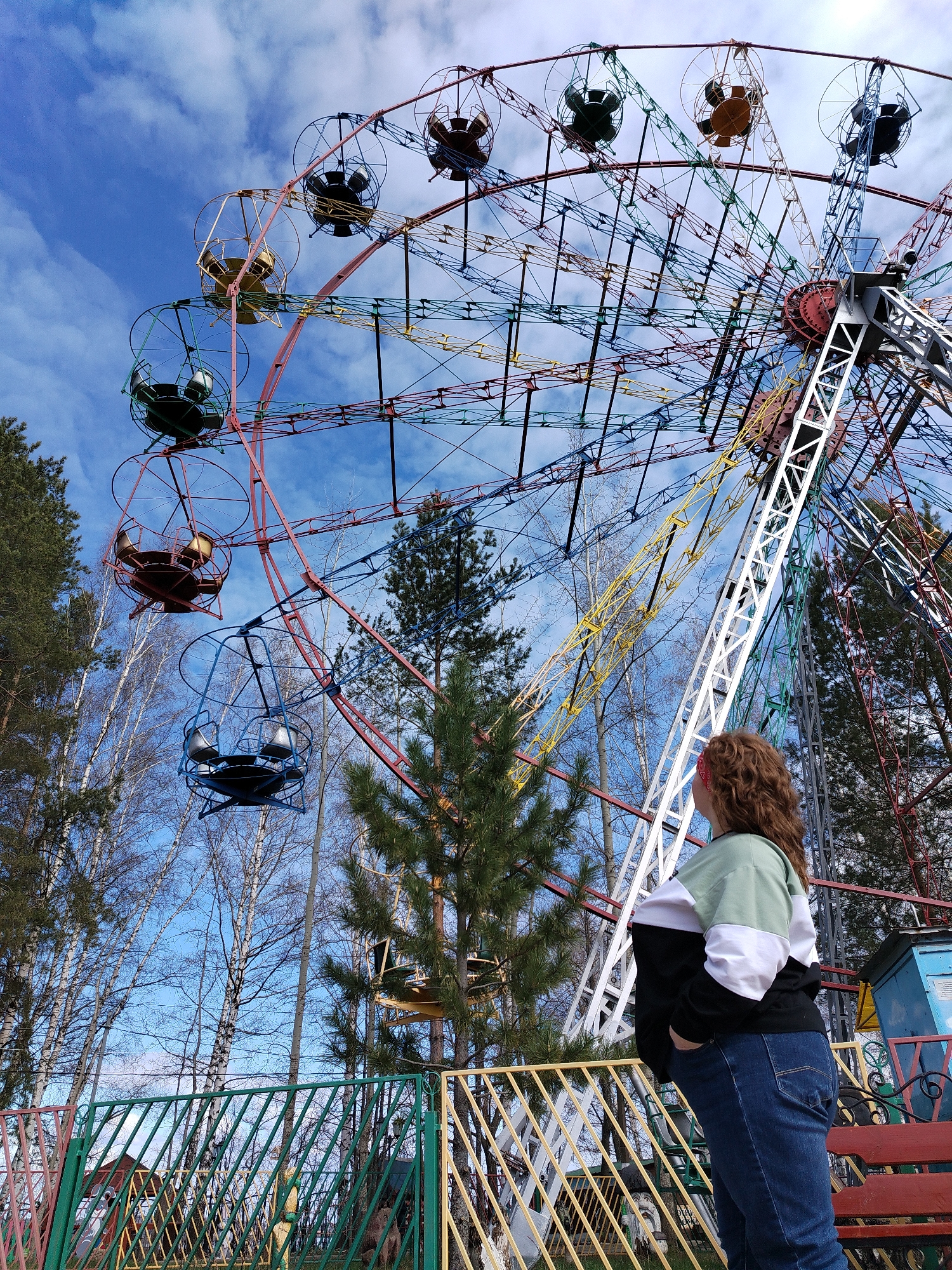 A woman with curly brown hair, wearing jeans and a black, white, and green top, stands with her hand in her pocket, looking up at a large Ferris wheel. The Ferris wheel is multi-colored, with red, blue, yellow, and green supports and carriages. The wheel is surrounded by trees, some bare and some evergreen, under a partly cloudy blue sky. The woman is standing behind a colorful metal fence with green, yellow, and red posts. A bench is visible behind the fence. The photograph appears to be taken in an outdoor recreational area or park. The location is Chaikovsky, Russia.