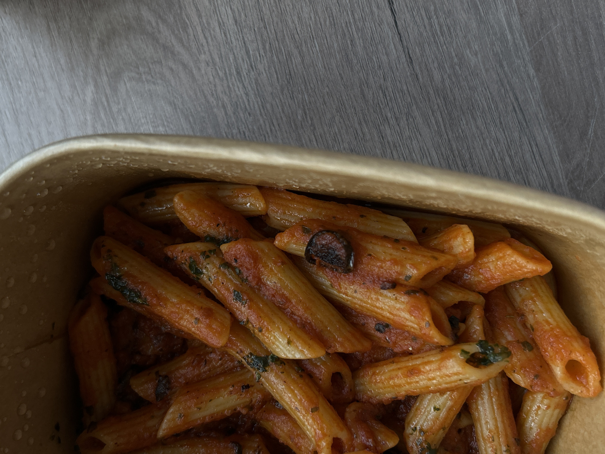 The image shows a container filled with penne pasta tossed in tomato sauce with herbs, likely basil. A dark, circular object, possibly a mushroom slice, is visible within the pasta. The container appears to be made of cardboard. The background is a gray, wood-grain surface. There is no indication of the location of Sofia, Bulgaria. There are no people present in the image.