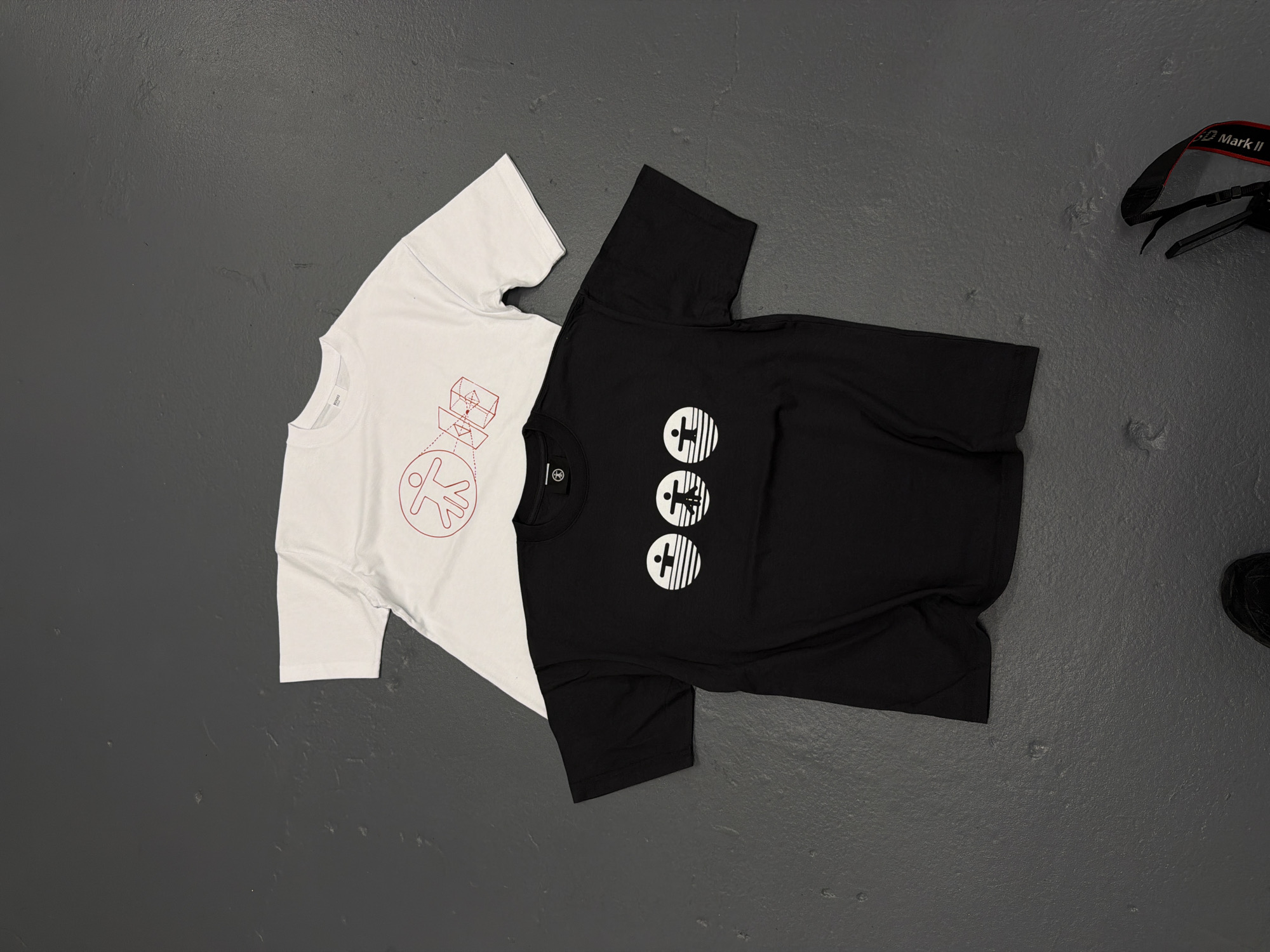 The image shows two t-shirts, one white and one black, lying on a gray surface. The white t-shirt has a red geometric design on it, while the black t-shirt has three white circular designs stacked vertically. In the upper right corner, there is part of a camera strap, with "Mark II" visible on it. In the lower right corner, part of a black shoe is visible. The location cannot be determined from the image.