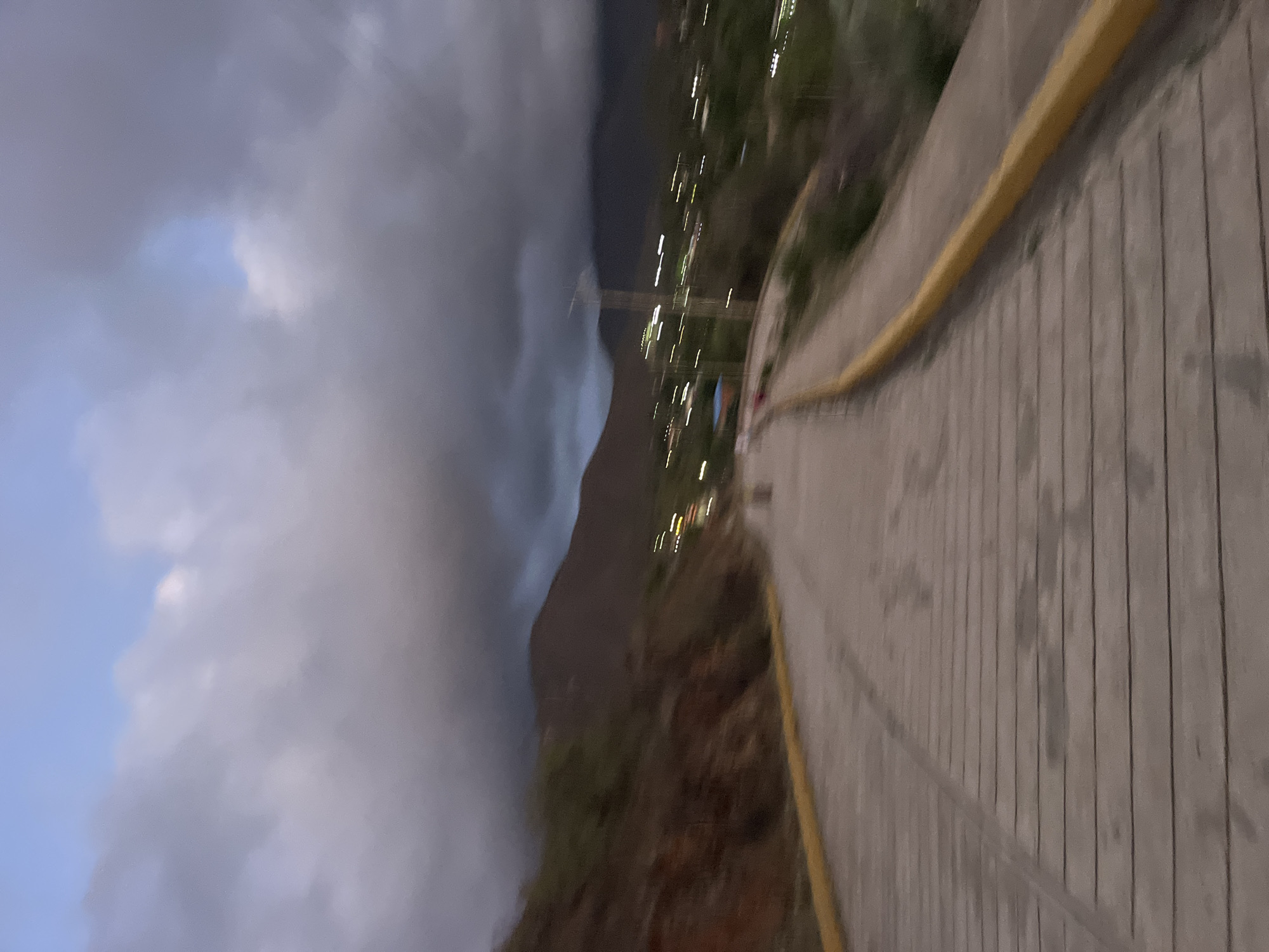 The image depicts a blurred view of a paved road with a yellow curb, leading uphill towards a town nestled at the base of a mountain range. The sky is overcast with dramatic clouds, hinting at twilight or an approaching storm. Streaks of light suggest distant streetlights or vehicle headlights. The terrain on the left is a steep, rocky slope covered with vegetation, while the right side features more greenery and the blurred lights of the town. There are no discernible people or specific objects that clearly identify the location within Santa Ana, Venezuela, but the mountainous backdrop and coastal-like vegetation are consistent with the region.