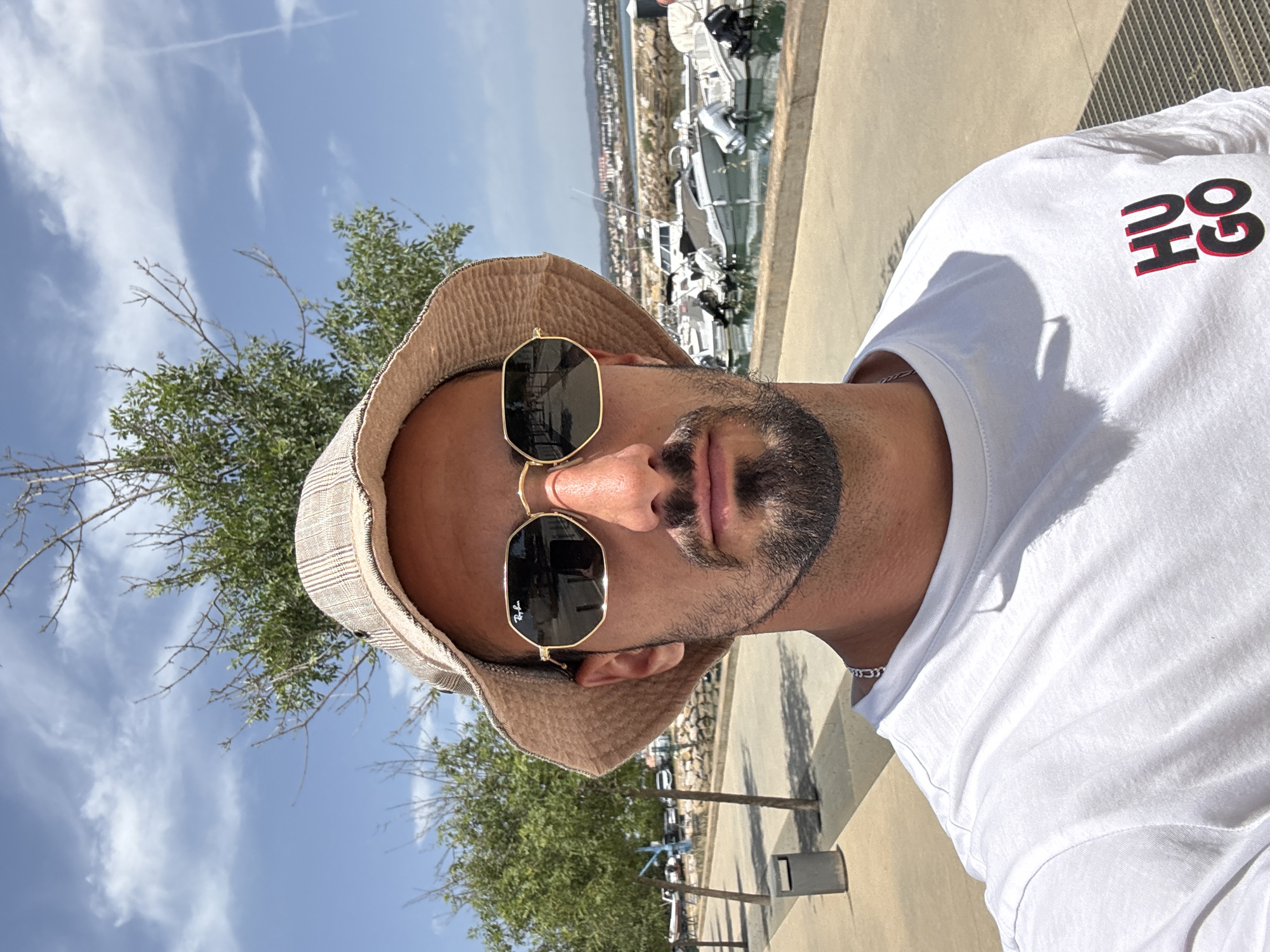 A person with a beard and sunglasses is wearing a bucket hat and a white t-shirt with upside-down red and black lettering, standing outdoors. The background includes trees, a blue sky with contrails, buildings, and boats in the distance. The setting appears to be a coastal or waterfront area. The location is Unknown, Unknown.