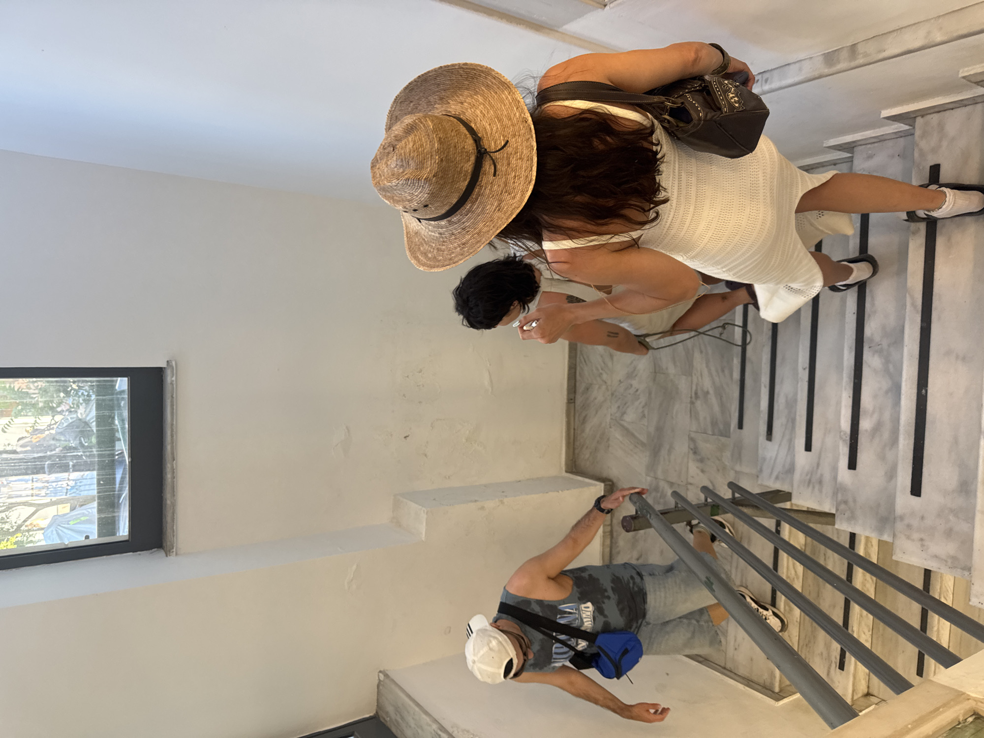 Three individuals are ascending a marble staircase. The person in the foreground, viewed from behind, wears a white knit dress and a straw hat. To their left, a person with dark hair in a white top is also ascending. Further ahead and to the left, a man wearing a white baseball cap, a tank top with a graphic print, denim shorts, and a blue fanny pack is descending the stairs. The staircase has grey metal railings and black strips on the edges of the steps. The walls are white and appear to be lightly textured. A window with a view of greenery and some obscured structures is visible at the top of the staircase. The lighting suggests an indoor or shaded outdoor environment. This scene is located in Chania, Greece, as indicated by the context provided.