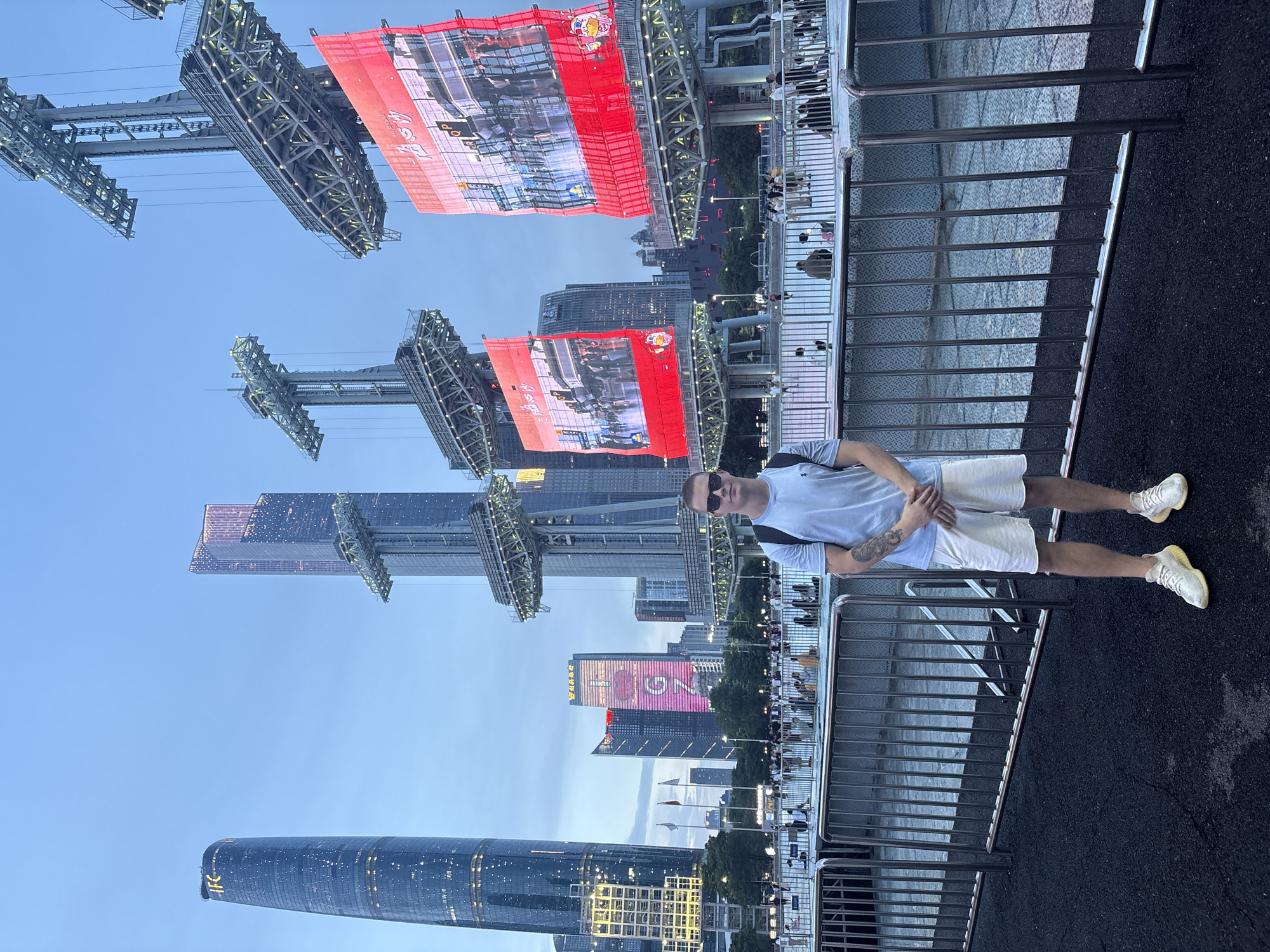 In Tianhe District, China, a young Caucasian male stands facing forward, positioned in the lower-center of the frame. He is wearing a light blue t-shirt, white shorts, and white athletic shoes. He has short dark hair and is wearing sunglasses. His arms are crossed in front of him. The background features a cityscape at dusk, with several modern skyscrapers illuminated against a twilight sky. Large, red digital screens are suspended from metallic structures, displaying images. A metal fence runs horizontally across the mid-ground, separating the viewer from a crowd of people gathered in the distance. The ground in the foreground is dark asphalt.