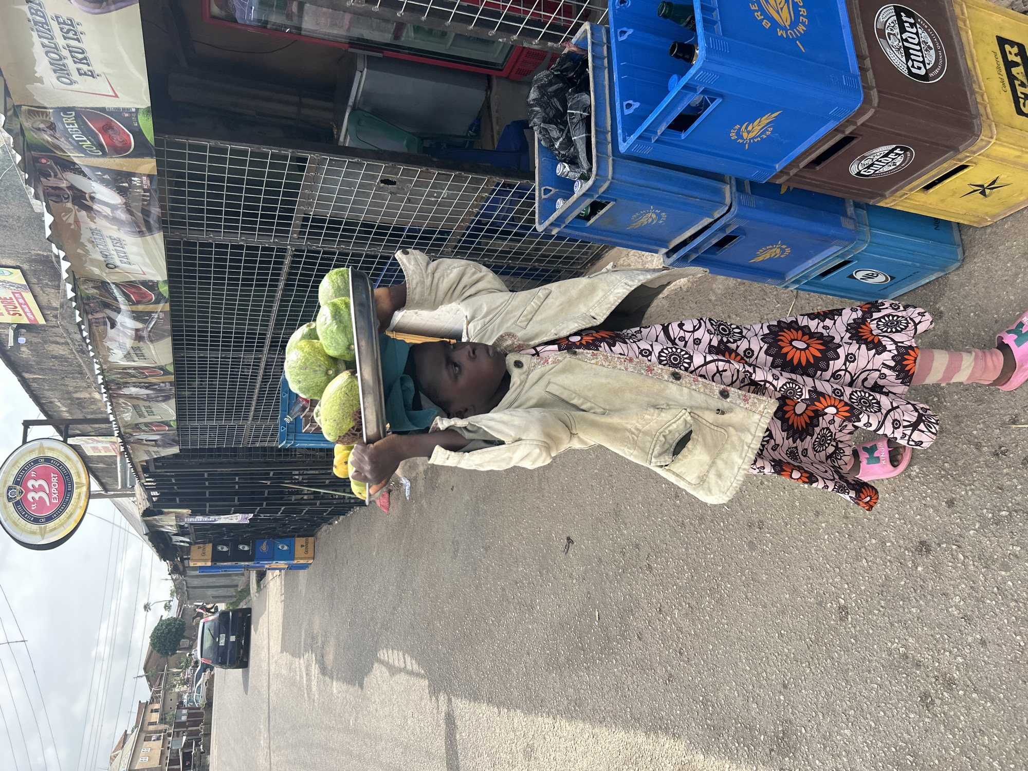 The image shows a child carrying a tray of green fruits on their head, walking on a paved road in what appears to be Jos, Nigeria. The child wears a floral print dress, a light-colored jacket, striped socks, and pink sandals. A stack of blue, brown, and yellow crates is visible alongside the road. Above the road, there are shops with awnings and signs, including one with "33 Export" on it. There are buildings and trees in the background, and a dark car is visible on the street. The overall scene is of a child working as a street vendor in an urban environment.