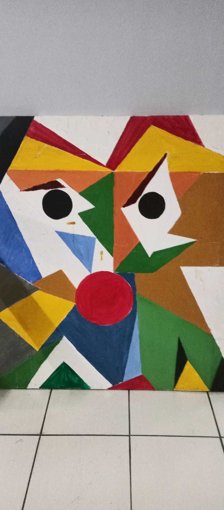 The image shows a close-up, vertically oriented view of a cubist painting of a clown's face. The painting is composed of various geometric shapes and bold colors, including red, yellow, blue, green, orange, white, brown, and black. The clown has two large black circular eyes with white angular shapes surrounding them, forming eyebrows and eye sockets. A prominent red circle sits in the center of the face, representing the nose. The background of the painting is textured white. Below the painting, a tiled floor with light-colored square tiles and dark gray grout lines is visible. The overall lighting is even, with no distinct shadows. The image was taken indoors, likely on a flat surface. The image provided does not contain any information to determine its location in the city of South Jakarta, Indonesia.