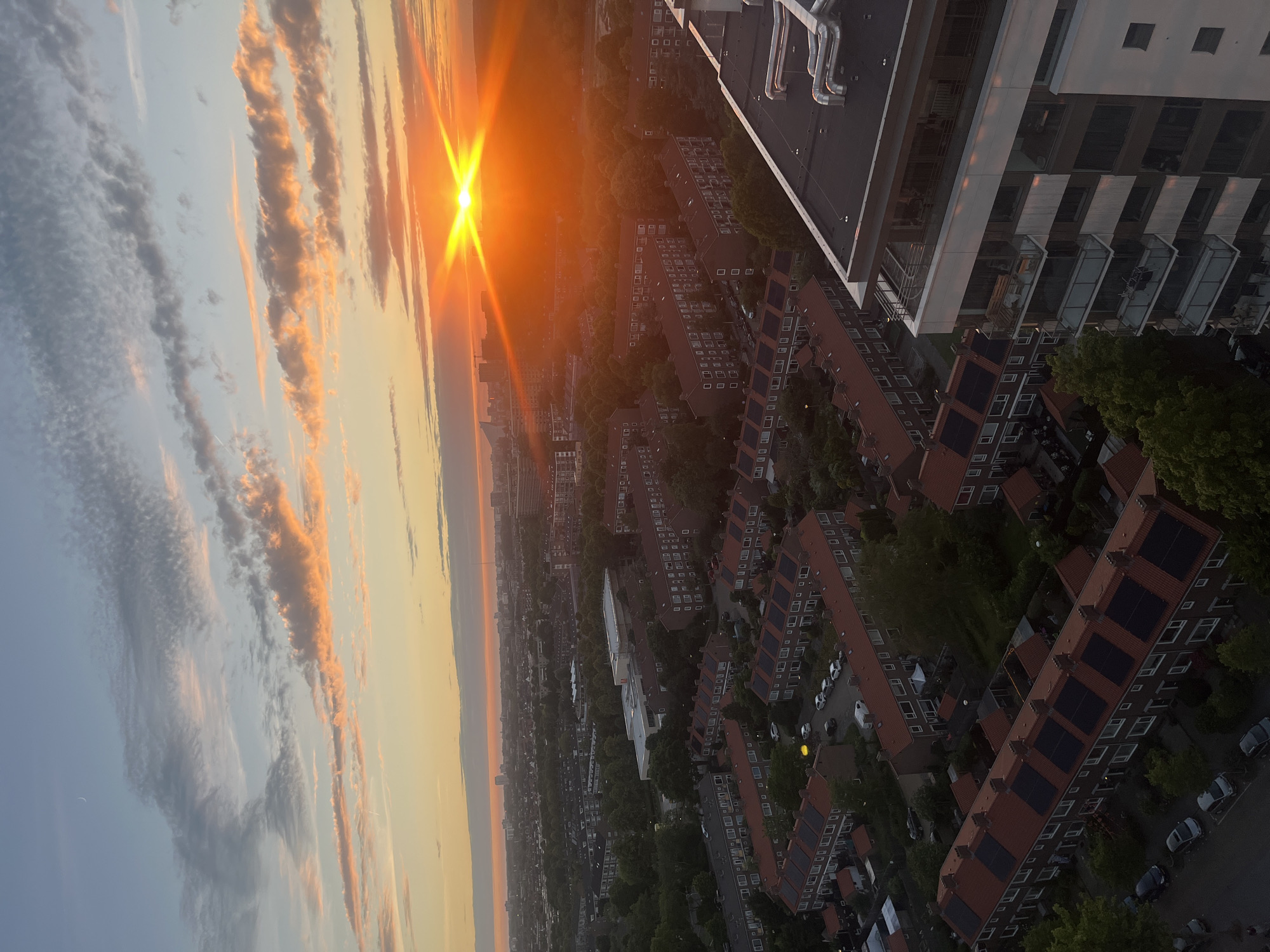 The image is an aerial view of a city at sunrise. The sky is filled with clouds that are illuminated by the rising sun, creating a colorful and dramatic effect. The sun is a bright orange orb with distinct rays of light emanating from it, creating a starburst effect. Below the sky, a dense urban landscape stretches out, characterized by rows of red-tiled houses with solar panels on their roofs, interspersed with patches of green trees. A modern apartment building with balconies is visible in the foreground on the right side of the image. The overall scene depicts the beginning of a new day in a residential area of a city.