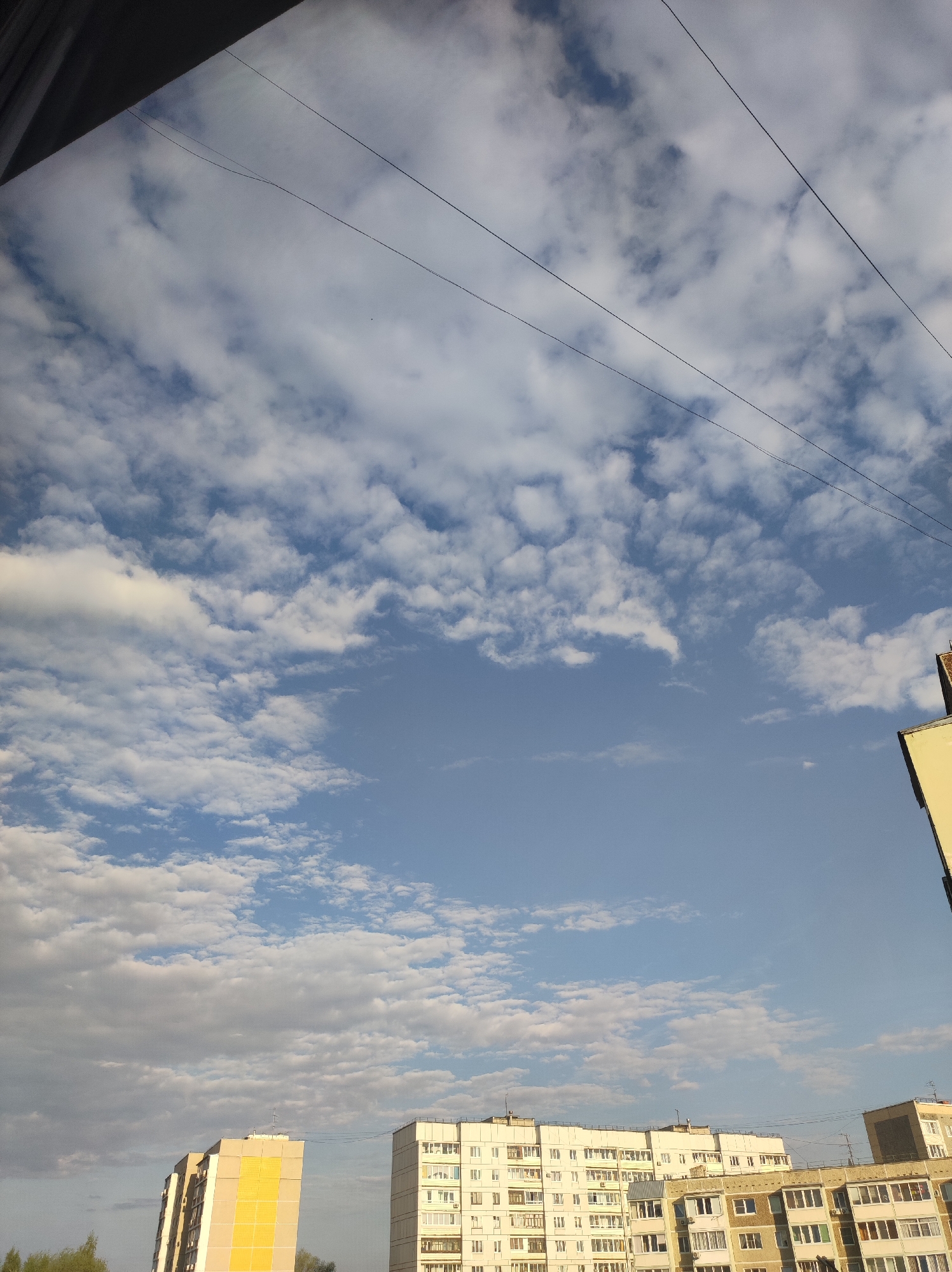 The image shows a view of the sky over Chaikovsky, Russia. The sky is a mix of blue and clouds. In the foreground are several apartment buildings. There are power lines running across the top of the image.