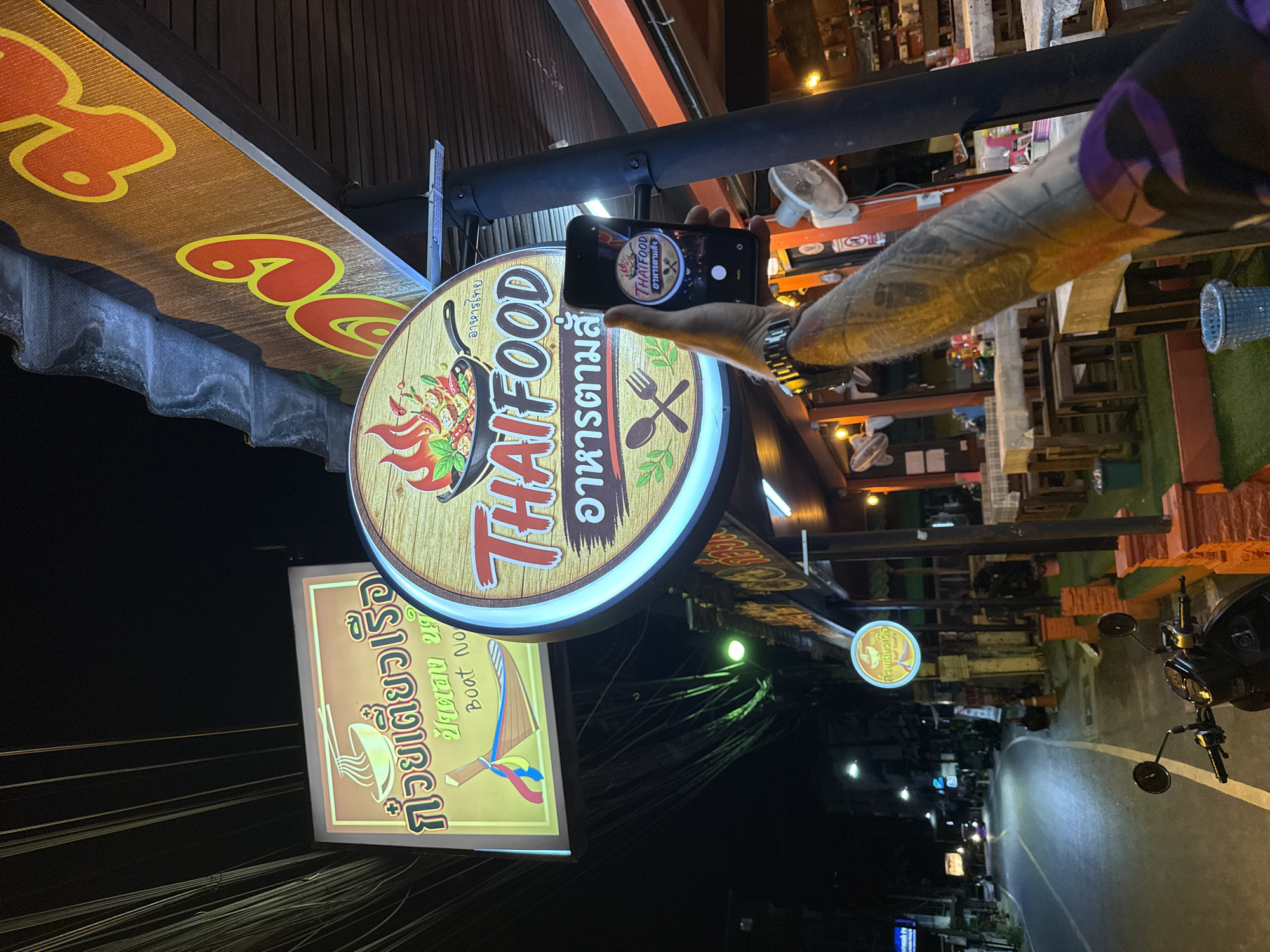 In an outdoor urban setting at night, a person's arm, adorned with extensive tattoos, is prominently featured in the foreground, holding up a smartphone to capture an image. The phone screen displays a close-up of a circular sign with the text "ThaiFood" and Thai script above and below, featuring a wok with flames and food. The main scene behind the arm shows a street lined with restaurants. A large, illuminated circular sign for "ThaiFood" hangs from the awning of one establishment, with Thai script above and below. To the left, a rectangular illuminated sign with Thai script and an illustration of a boat with noodles is also visible. The restaurants appear to be open, with tables and chairs set up on a green-tiled patio. The street is paved and illuminated by streetlights, with a motorcycle parked on the side. The overall atmosphere suggests a vibrant, late-night dining district in an Asian city.
