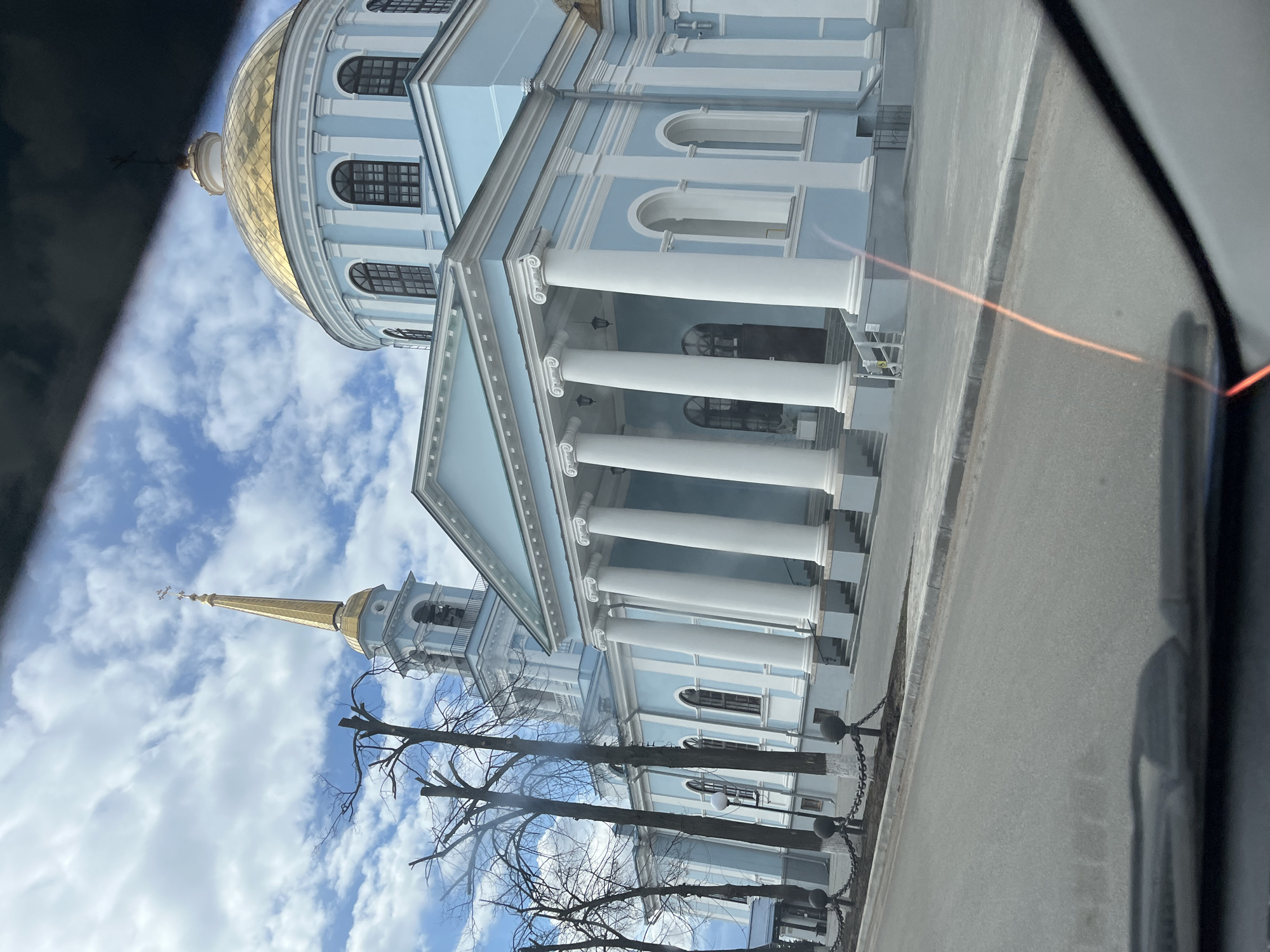 The image captures a light blue and white church or cathedral under a partly cloudy blue sky, viewed from inside a vehicle in Votkinsk, Russia. The prominent architectural style features a large, ornate golden dome on the main structure, and a taller, slender golden spire with a cross atop a bell tower to the left. The building's facade is adorned with white classical columns supporting a portico, and features arched windows.
In the foreground, bare deciduous trees with recently trimmed branches are visible. A grey asphalt road runs along the front of the building, separated by a curb and a chain fence with dark spherical bollards. On the right side of the frame, parts of a car's interior, likely a door frame and window, are visible, indicating the photo was taken from within a vehicle. A reddish-orange light streak, possibly a reflection or lens flare, cuts across the mid-ground.
The scene depicts a calm daytime setting with clear weather, suggesting a sunny day with scattered clouds. No specific human activity or events are visible. There is no discernible text within the image.