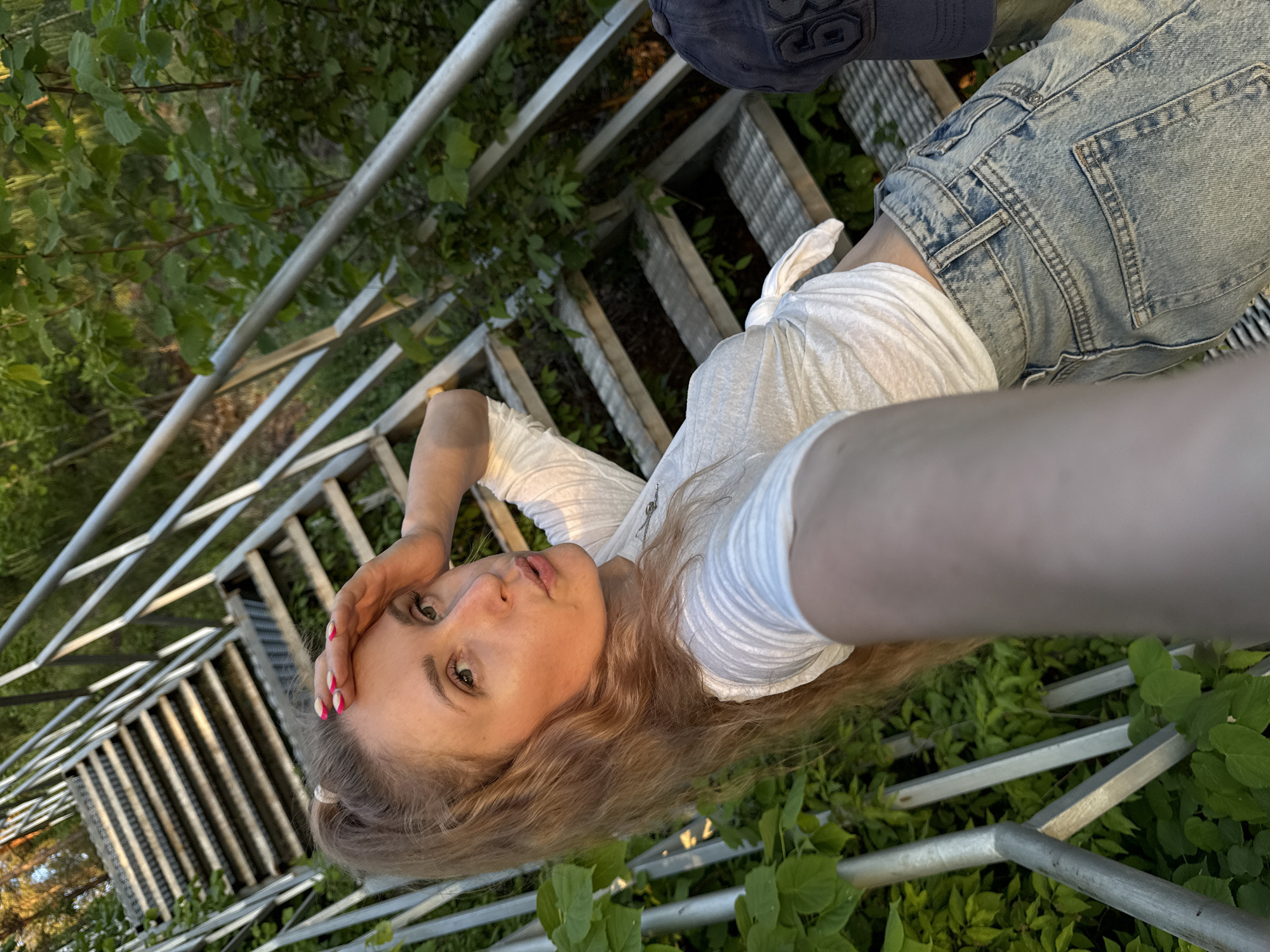 A young woman with reddish-blonde hair, wearing a white top and light-wash denim shorts, is positioned on a metal ladder or scaffolding structure. She is angled in the frame, with her head near the bottom and her legs towards the top. Her hand is near her face, and she is looking at the camera. The ladder is situated amidst greenery, likely foliage. The background is blurred, but there is a sense of being outdoors in a forested or garden setting. The lighting suggests it is late afternoon or early evening. The environment appears to be outdoors with lush greenery, possibly the city of Noviy, Russia.
