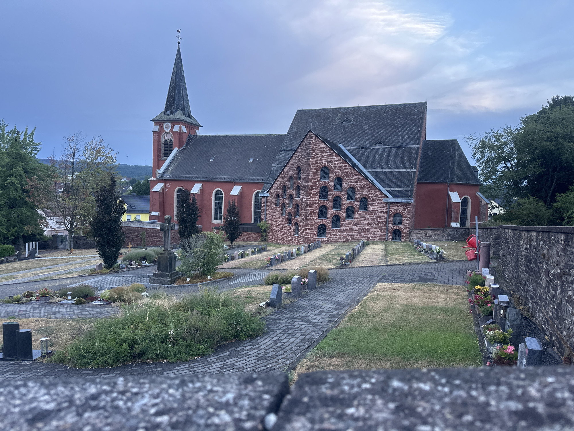The image depicts a cemetery adjacent to a red brick church in Wadern, Germany. The sky is overcast with a pale blue hue, suggesting either early morning or late evening. The church features a tall, pointed steeple with a cross at its apex and a clock face. The main body of the church is constructed of red brick, with arched windows visible. An additional section of the church is built with reddish-brown brick and features a repeating pattern of arched openings. The cemetery is laid out with rows of gravestones and small floral arrangements. Paved pathways wind through the cemetery. A stone cross stands prominently in the foreground of the cemetery. Greenery, including trees and bushes, is present throughout the scene. A stone wall borders the right side of the cemetery. No people are visible in the image.